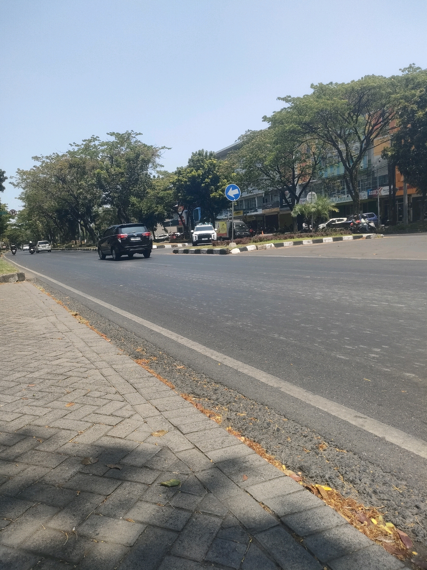 The image shows a street scene in Surabaya, Indonesia, on a clear, sunny day. A wide, paved road occupies the majority of the frame, with a sidewalk made of interlocking grey pavers on the left. The road has a white dividing line. Several vehicles are visible on the road, including a black SUV in the foreground, a white SUV further back, and several motorcycles. Trees line the side of the road, with lush green foliage. Behind the trees, a multi-story building with a colorful facade is visible. The building appears to be a commercial establishment, with signs for various businesses. The sidewalk has some fallen leaves scattered on it, and the edge of the road is lined with a strip of dark material. The sky is a clear, pale blue.