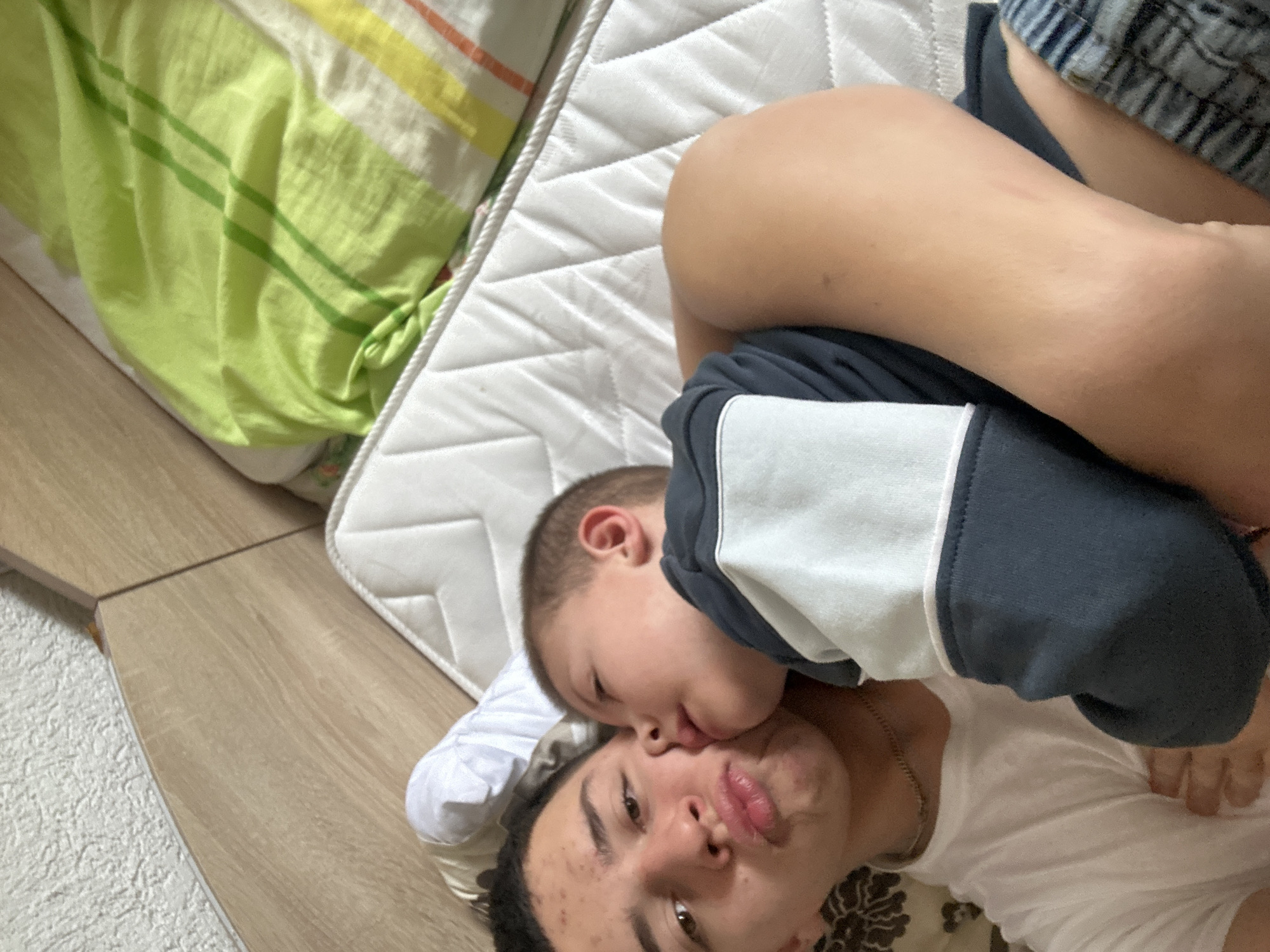 The image displays an intimate moment between two individuals, an adult and a child, lying down indoors. The adult, positioned in the foreground, lies on their back, looking towards the camera with slightly puckered lips. They have dark, curly hair, a light complexion with some blemishes, and wear a plain white t-shirt and a thin gold chain.
Cuddled closely on top of the adult, the child has short, light brown hair and is wearing a dark blue and white long-sleeved top. The child's face is pressed against the adult's cheek, appearing to give a tender kiss or snuggle, which conveys a strong sense of affection and closeness.
They are resting on a white, quilted mattress, which is placed on a wooden floor or platform. A bright green and striped blanket or sheet is partially visible in the upper left portion of the frame. To the left of the mattress, a section of a white, textured rug can be seen beside the wooden floor. The setting suggests an indoor environment, possibly a bedroom or casual living space. The lighting is bright, indicating it is likely daytime. No visible text is present in the image. This media file is provided with the location context of Zlatograd, Bulgaria.