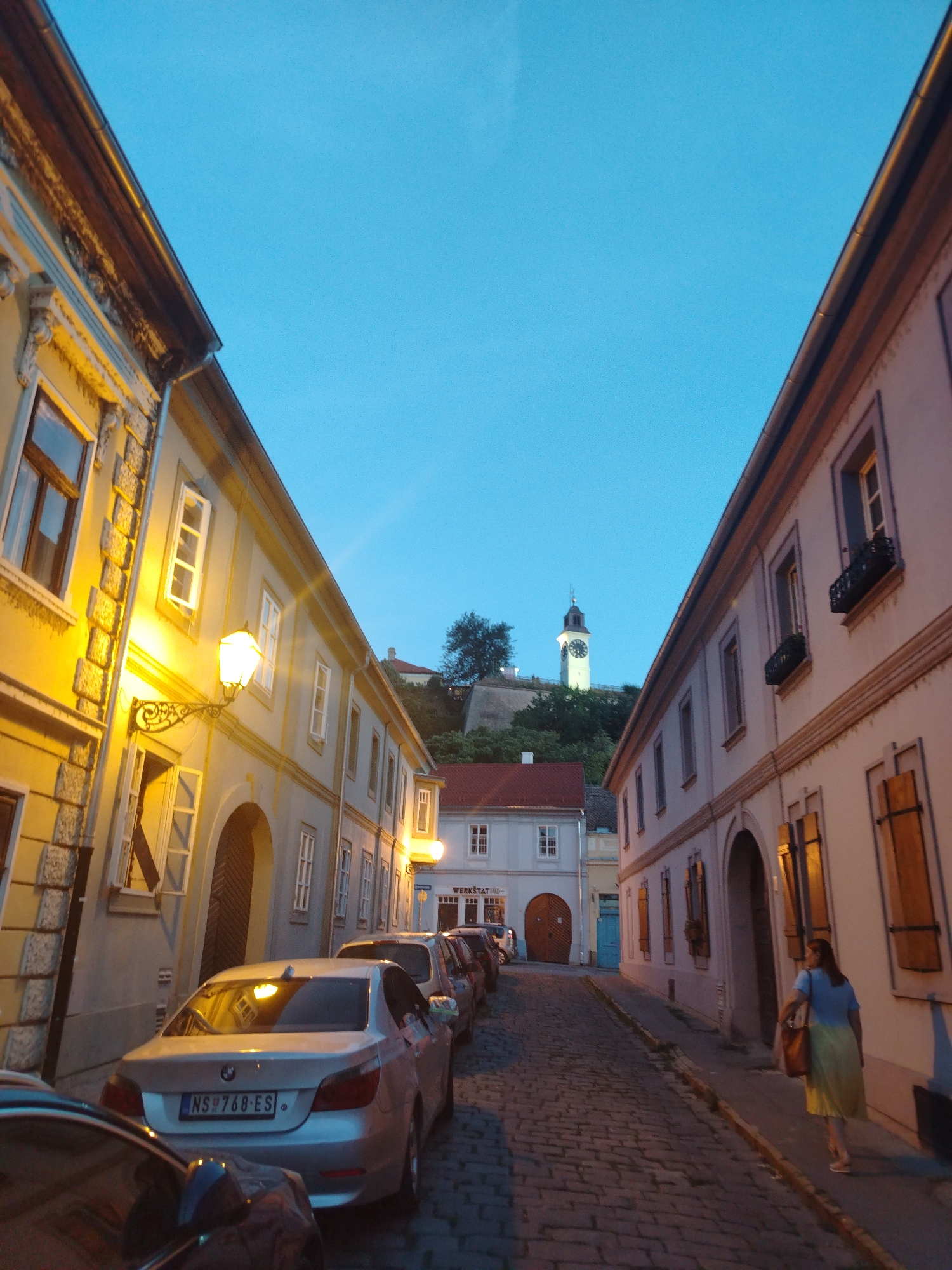 The image captures a street scene in Petrovaradin, Serbia, at dusk. The perspective is looking up a cobblestone street lined with old, multi-story buildings. The sky is a deep twilight blue. On the left, a building features a yellow facade with ornate architectural details and a glowing lantern. On the right, a building with a light-colored facade has closed wooden shutters and a woman in a blue shirt and yellow skirt walks away from the camera. Several cars are parked along the street, including a silver BMW sedan with a license plate reading "NS 768-ES". In the distance, a clock tower with a white facade and a dark roof stands prominently on a hill amidst trees, a characteristic landmark of Petrovaradin Fortress.