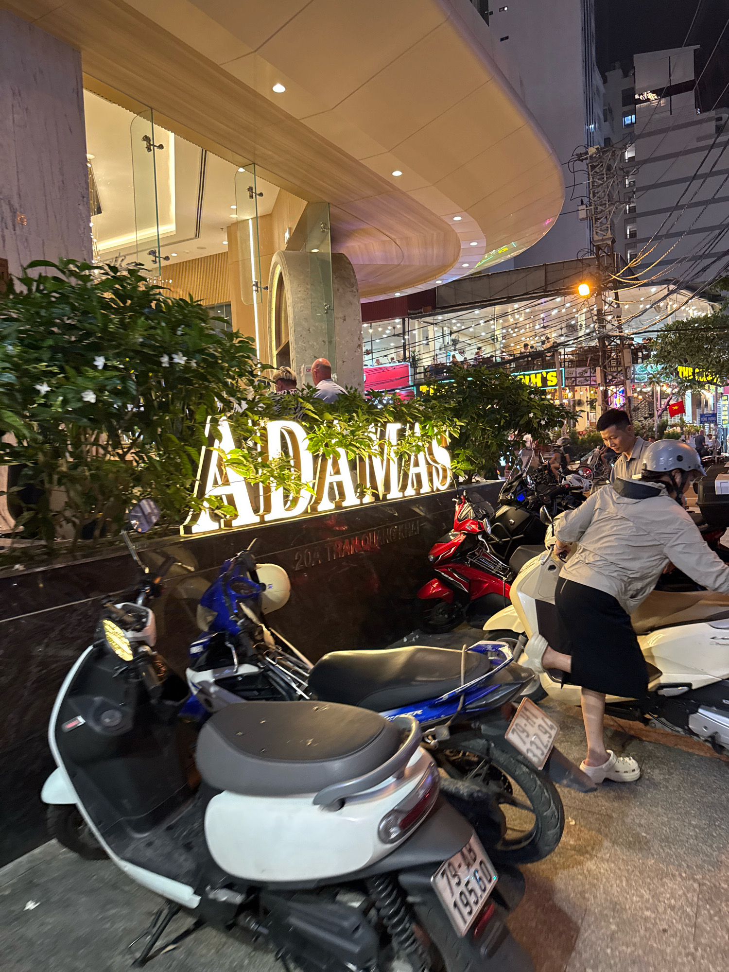 This image captures a street scene in Nha Trang, Vietnam, at night. A large, illuminated sign with white lettering reads "ADAMAS" in the center of the frame, positioned above a dark, polished surface and surrounded by lush green foliage. The building behind the sign features a modern, curved architectural design with warm lighting. In the foreground, several parked motorcycles are visible, with one white scooter and a blue and white scooter most prominent. A person wearing a light-colored jacket, black skirt, and white Crocs is interacting with a motorcycle, appearing to be preparing to mount it. Another person, a male, is standing nearby, looking towards the motorcycles. The background reveals a street with more illuminated buildings, overhead electrical wires, and streetlights, indicating a commercial or entertainment area. The overall atmosphere suggests a bustling urban environment at dusk or night.