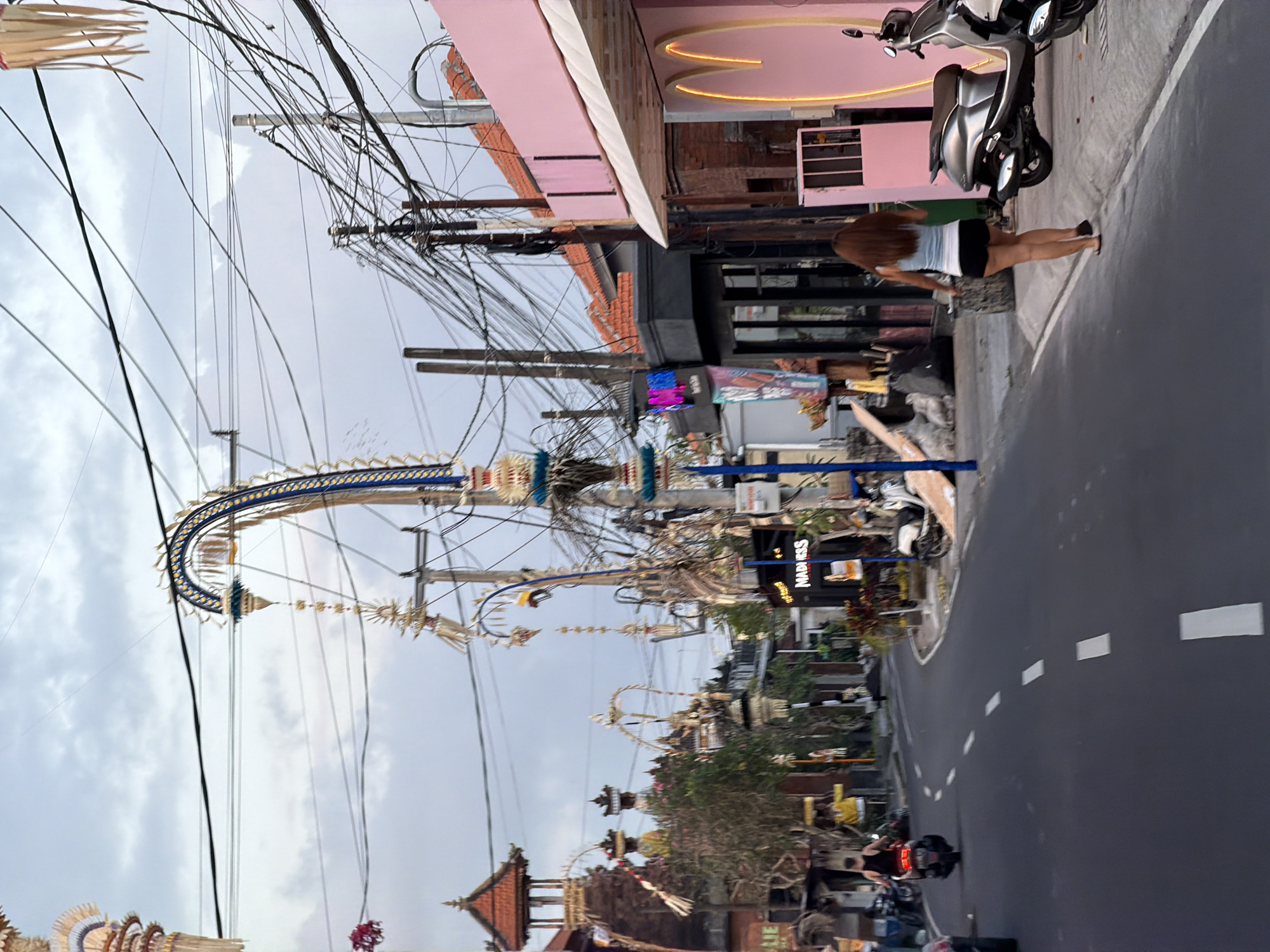 The media file captures a street scene in Mengwi, Indonesia, likely during the daytime with overcast skies. The image is oriented vertically, showing a busy road lined with buildings and adorned with numerous decorative elements.
Prominently featured are intricate, arching structures that appear to be part of a festive or ceremonial display, possibly related to a local festival or tradition. These decorations are suspended above the street, intertwined with a dense network of electrical wires, creating a visually complex overhead environment.
On the right side of the road, a modern silver scooter is parked near a building with a pink facade. A woman with long brown hair, wearing shorts and a top, stands facing away from the camera, appearing to be waiting or observing. Further down the road, another scooter with a rider can be seen, their brake lights illuminated, suggesting movement.
Buildings along the street exhibit a mix of architectural styles, from traditional tiled roofs to more contemporary designs with colorful signage. One sign clearly visible reads "MADNESS." The street itself is paved and marked with white dashed lines, indicating a road for vehicles.
The overall impression is one of a vibrant, somewhat chaotic urban environment, possibly during a special occasion that involves elaborate decorations and a significant presence of motorcycles, common modes of transport in the region. The cloudy sky suggests it might be humid or on the verge of rain.