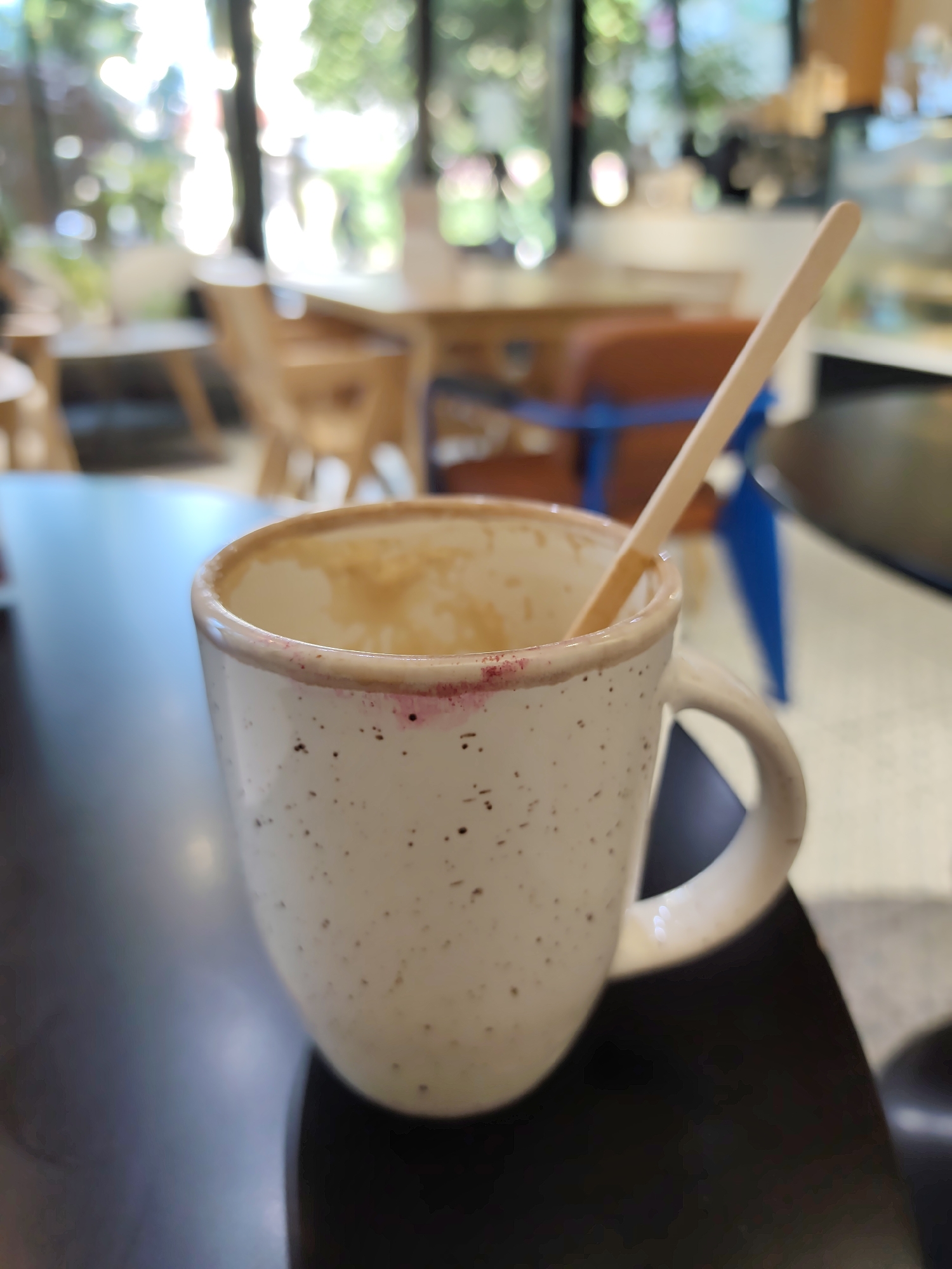 A close-up, shallow depth-of-field shot focuses on a speckled white ceramic mug sitting on a dark surface. A light-colored wooden stirrer extends from the mug, which contains a small amount of residual brownish liquid. A faint pink stain, possibly lipstick, is visible on the rim of the mug. The background is blurred, showing the interior of what appears to be a cafe or restaurant with tables, chairs, and natural light filtering in from windows. The overall impression is of a recently used beverage container in a public space.