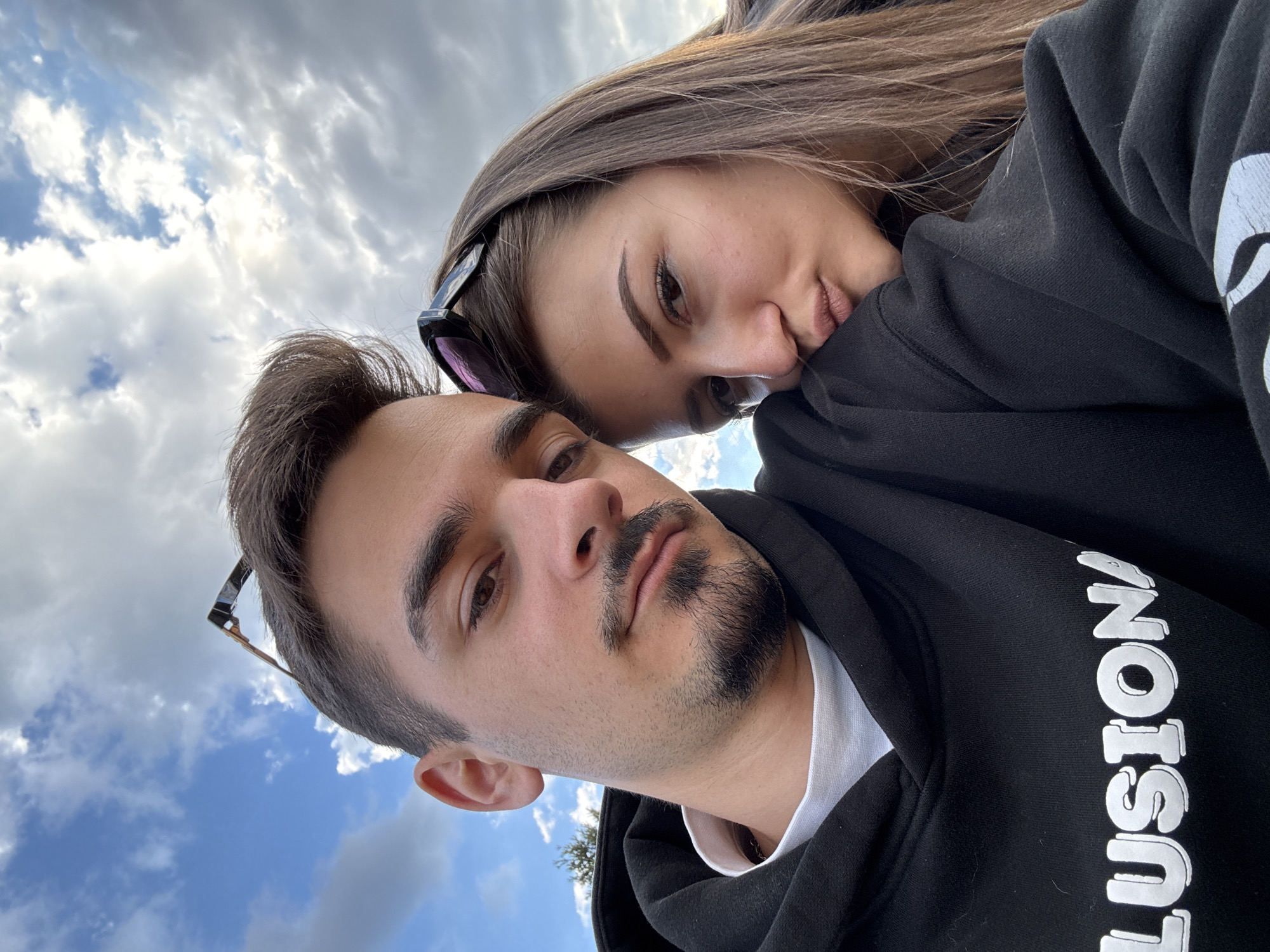 A close-up selfie shows a young couple outdoors, likely in Sofia, Bulgaria, given the context. The man, on the left, has short dark hair, a mustache, and a goatee. He is wearing a black hoodie with white text that reads "LUSION" and a white t-shirt underneath. Sunglasses are perched on his head. To his right, the woman rests her head on his shoulder. She has long, light brown hair and is wearing a black hoodie. Her lips are slightly parted in a pout. Behind them, a cloudy blue sky suggests it is daytime. The overall impression is one of a casual, affectionate moment captured outdoors.