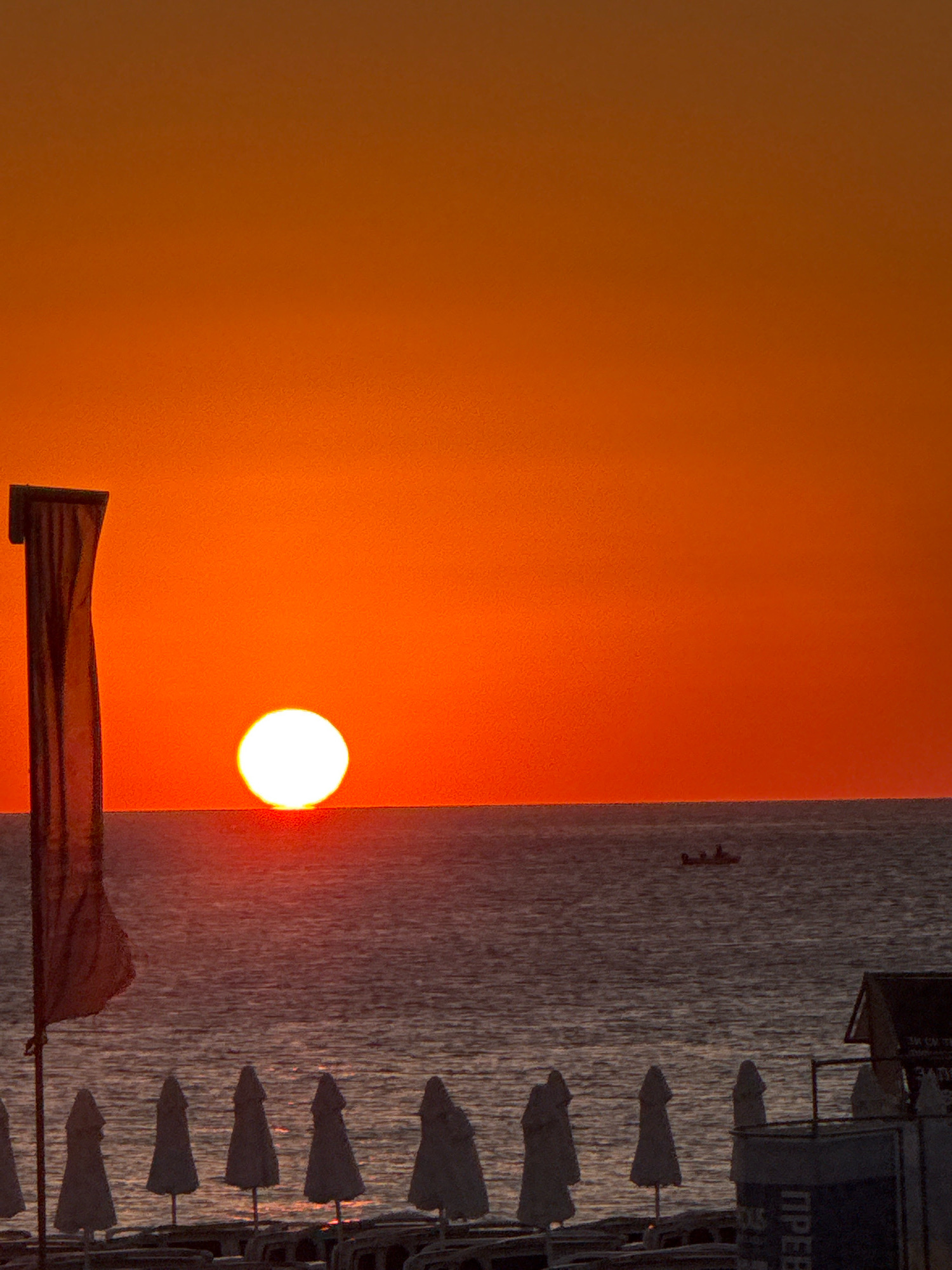 This image depicts a serene sunset over the Black Sea, viewed from a beach likely located in Sozopol, Bulgaria, given the context provided. The dominant colors are vibrant oranges and reds, saturating the sky and reflecting on the water's surface.
In the foreground, a row of closed beach umbrellas stands in neat formation, suggesting the beach is not actively in use. Beyond the umbrellas, the gentle waves of the sea stretch towards the horizon. A single, small boat with silhouetted figures can be seen in the distance, gently navigating the water. To the left of the frame, a tall, dark flag or banner stands prominently, its fabric hanging loosely. On the far right, a small, rustic beach structure or kiosk is partially visible, with some signage that is too blurry to read. The sun, a bright white orb, is positioned just above the horizon, casting its warm glow across the scene. The overall atmosphere is tranquil and picturesque, capturing a moment of natural beauty at the end of the day.