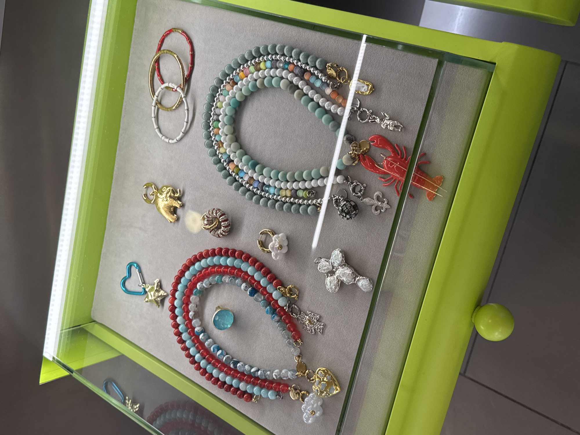 The image displays a collection of jewelry displayed inside a bright green-framed glass case. The background within the case is a light gray. The jewelry consists of several beaded necklaces, some arranged in loops, and various charms or pendants scattered throughout. A red lobster charm is visible. There are also rings, gold-colored charms, and other decorative items. The location is Dorozhnyi, Russia.