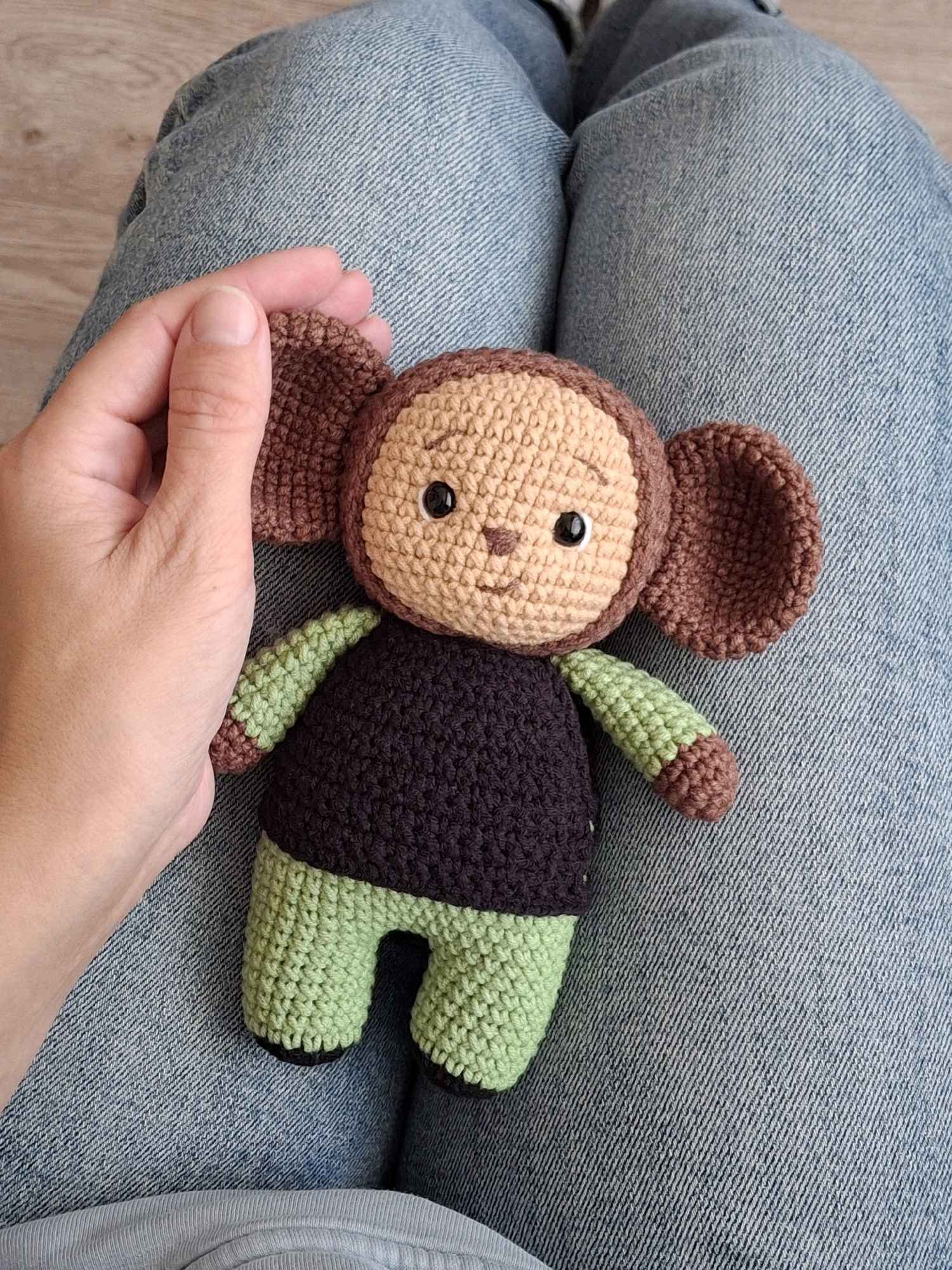 A hand is holding a crocheted doll, which appears to be a monkey, on top of a pair of denim jeans. The doll has a tan face with black eyes and a stitched smile, brown ears, a dark purple body, and green arms and legs with brown tips. The jeans are a light blue denim with visible stitching and texture. The background shows a light-colored wooden floor and more of the denim jeans. The scene does not contain any identifiable landmarks or features that would indicate its location in the city of Chaikovsky, Russia.