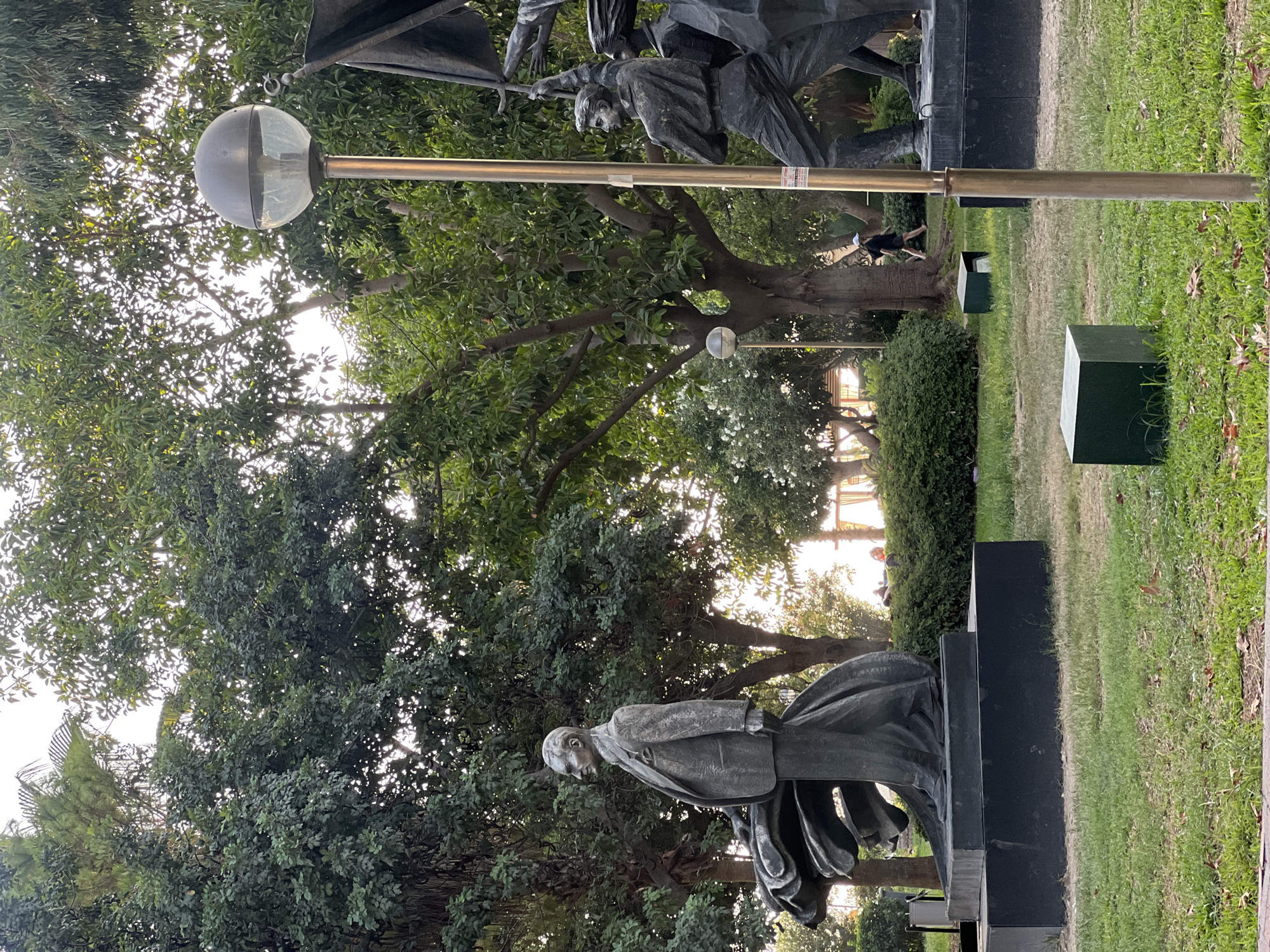 This image captures a statue of Mustafa Kemal Atatürk in a park, likely located in Alanya, Turkey, as suggested by the visual context. The statue depicts Atatürk in a suit, standing to the left of the frame and looking towards the left. To the right of the statue, a lamppost with a spherical shade is visible. Further to the right, there is another statue featuring several figures holding flags, suggesting a theme of national pride or historical significance. The park is characterized by lush green grass, mature trees with dense foliage, and sunlight filtering through the leaves, creating a dappled lighting effect. In the background, a person is walking in the distance, and there are indications of public seating areas, possibly benches. The overall atmosphere is serene, with elements of nature and historical monumentality coexisting.