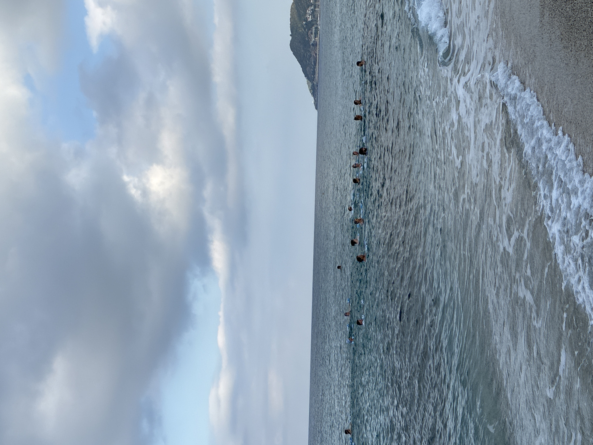 A group of approximately 15 individuals are swimming in the sea off the coast of Alanya, Turkey. The water is calm with small waves lapping at the shore in the foreground. The sky is partly cloudy with a mix of grey and white clouds. In the background, a green, hilly peninsula with some buildings and a lighthouse is visible. The people are scattered across the water, some with their heads fully above the surface, others with only their heads visible. They appear to be engaged in leisure activity or perhaps a swimming lesson.