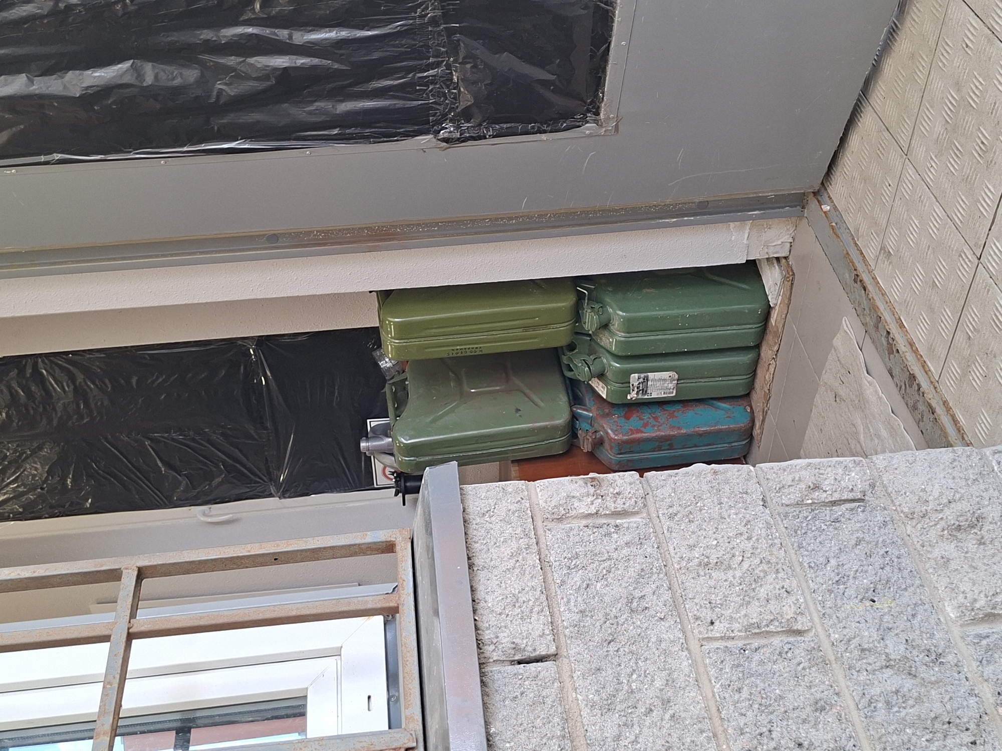 This image captures a utilitarian outdoor or semi-enclosed storage space, situated in what is identified as Saint Petersburg, Russia. The primary subjects are a collection of stacked jerrycans, commonly used for fuel or other liquids.
At least five jerrycans are visible. Three are a distinct olive-green color: two are stacked on the left, and one is on the right. Below these are two older, more worn cans with a faded, greenish-blue hue, one prominently positioned on the bottom right. Many of the cans show signs of age, with the lower blue-green ones exhibiting considerable rust and paint deterioration.
A significant detail is the text on the top-left green jerrycan, which clearly displays "K 00 0704 S" and below it, "СДЕЛАНО В С," strongly indicating "СДЕЛАНО В СССР" (MADE IN USSR). Another green jerrycan on the top right has an unreadable white sticker.
Above the stored jerrycans, an opening or window is covered with black plastic sheeting, secured with tape, suggesting a temporary covering for insulation or to block visibility. This area is framed by gray metal panels, possibly part of a door or window frame. To the lower left, a white window frame with a rusty metal grate in front of it is partially visible.
The surrounding structures include a light-colored, textured tiled wall on the right side, featuring a raised diamond-plate pattern. In the foreground, a wall constructed from rough-hewn, light-colored stone or concrete blocks is visible. A rusted metal strip runs along the base of the tiled wall. No human activity is depicted; the scene exclusively focuses on the stored containers and their immediate environment. The lighting suggests it is daytime.