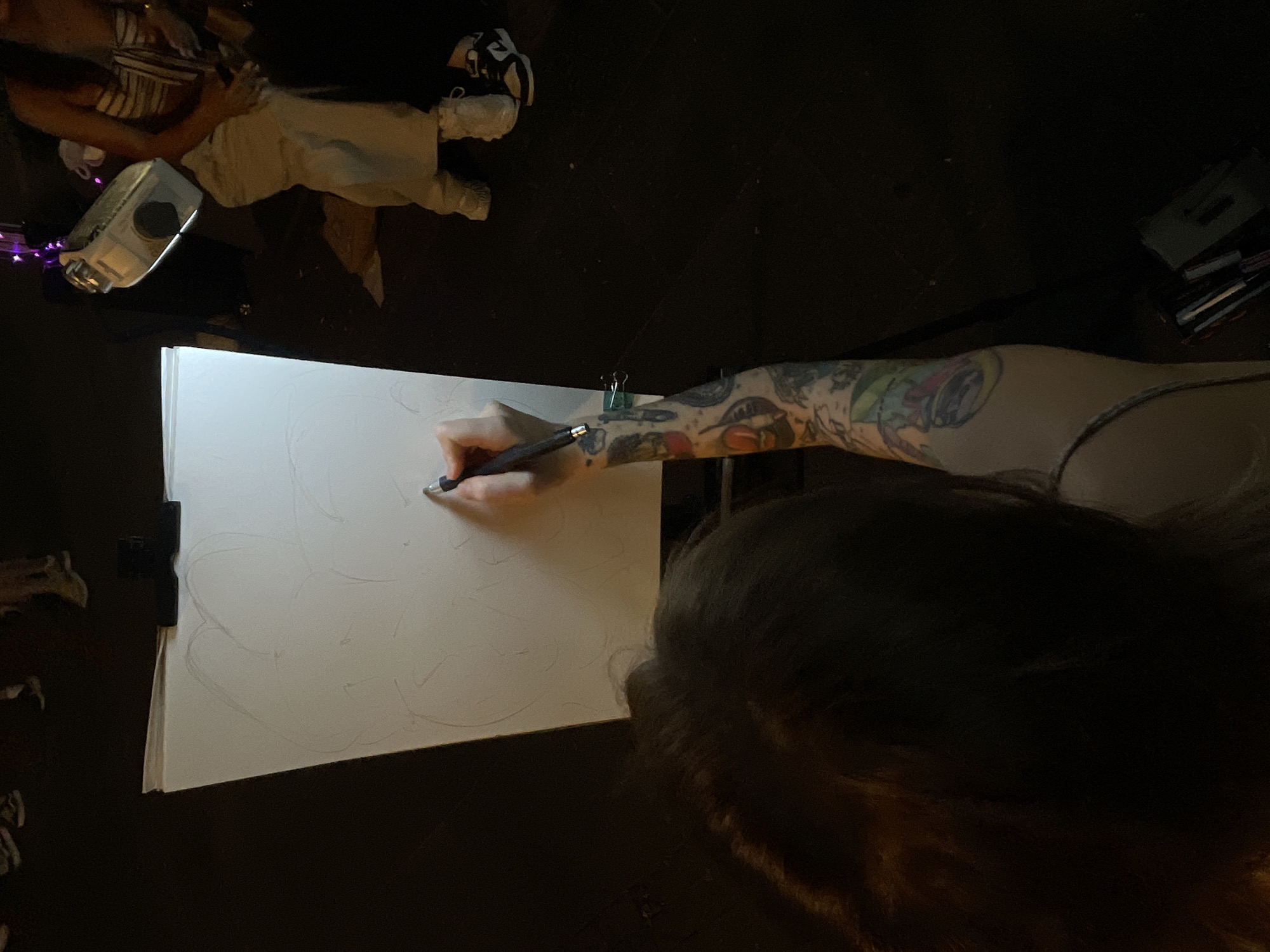 An artist with a tattooed arm sketches on a large canvas on an easel. The artist's face is obscured by shadow. In the background, other people are visible, including a person in light-colored pants and a striped top holding a white object with a black circular opening, possibly a portable speaker. The scene appears to be outdoors at night, with a dark ground surface, suggesting an urban environment such as a promenade or public square. The overall atmosphere is informal, potentially a street art event or an artist working in a public space. Without specific landmarks or context clues, pinpointing an exact location in Belgrade, Serbia, is not possible from the visual information alone.