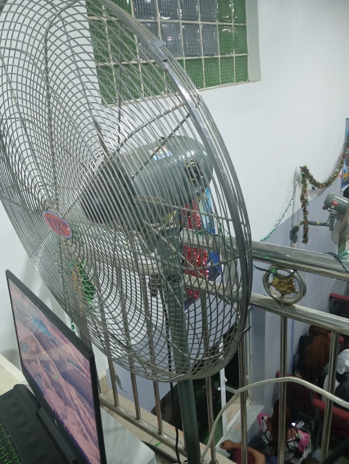 A metallic floor fan dominates the scene, its circular guard filling much of the frame. To the left, a computer monitor displays a scenic image, partially obscured by a dark keyboard. The fan stands near a staircase railing adorned with garland, suggesting a festive setting. Below the railing, a glimpse of people and chairs hints at activity on a lower level. A window with a grid pattern is visible in the background.