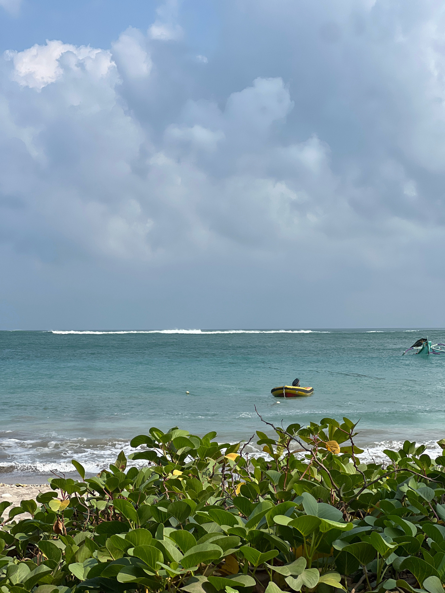 In Kuta, Indonesia, a view of the ocean is captured on an overcast day. The foreground is filled with lush green foliage, obscuring the sandy beach below. The ocean, a vibrant turquoise, features a line of white-capped waves breaking on the horizon. Two small boats are visible in the water: one yellow and black inflatable boat with an outboard motor, and another, larger boat to the right, partially submerged and painted green. The sky is a mix of light and dark gray clouds, with patches of brighter sky suggesting the sun is intermittently visible.