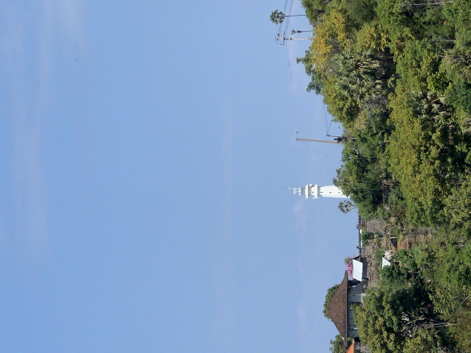 The image captures a coastal landscape in Abang, Indonesia, dominated by a clear blue sky. In the middle ground, a white lighthouse stands tall atop a hill, surrounded by lush green foliage and some scattered structures. One prominent building with a reddish-brown roof is visible to the left of the lighthouse. The hill is covered with a variety of trees and vegetation, with some rocky outcrops visible. Several utility poles with wires are also present on the hill, suggesting the presence of infrastructure. The foreground is densely packed with trees, creating a sense of depth. The scene appears to be a daytime view, with ample sunlight illuminating the landscape.