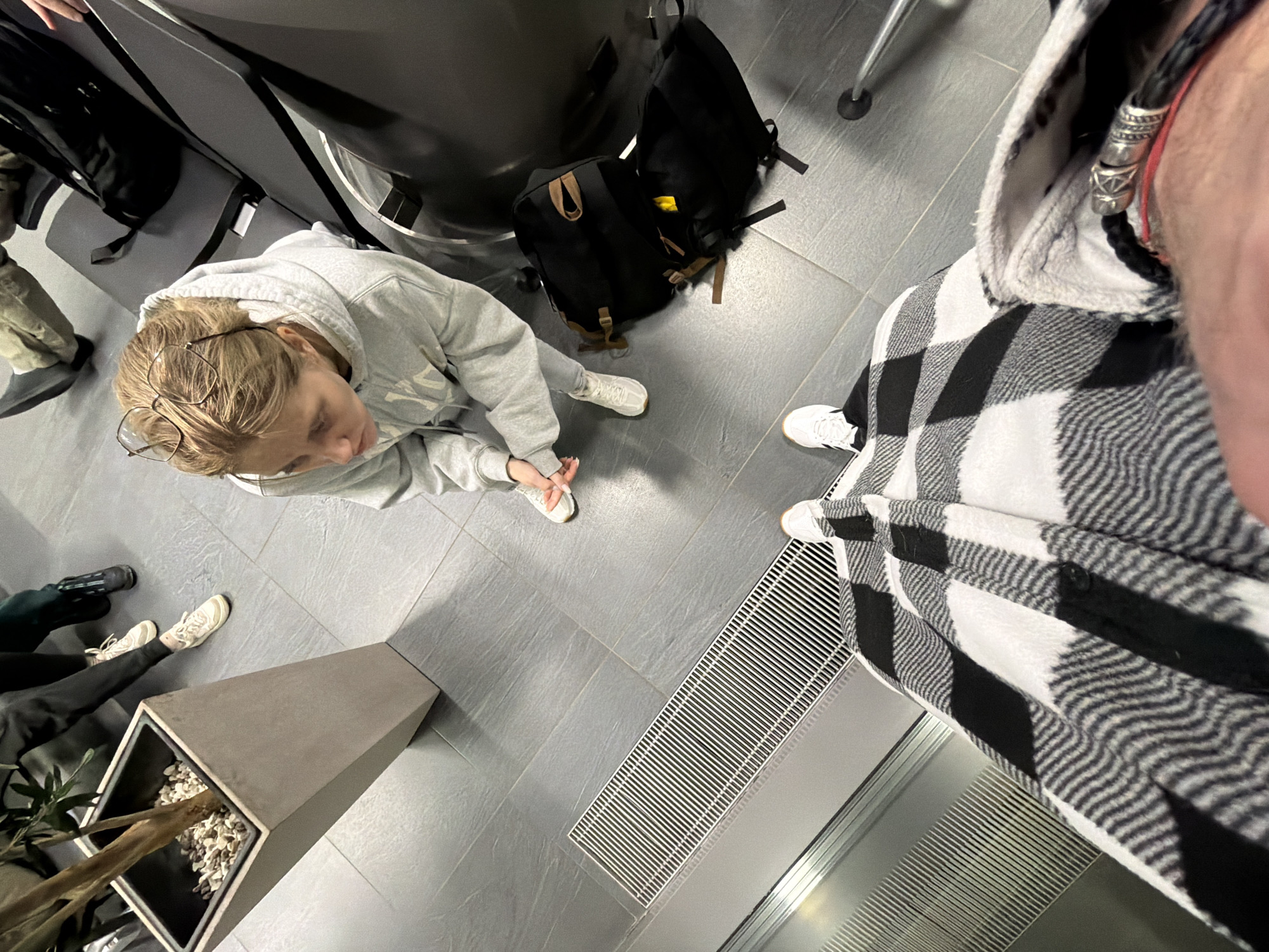 This image depicts an indoor public waiting area, possibly an airport or transportation terminal, located in Zarechnaya (Gamovskoe S/P), Russia. The scene is captured from a first-person perspective, looking down and forward from an individual standing on a metallic escalator or moving walkway. The photographer's lower body is visible, wearing white athletic shoes and black trousers, covered by a black and white buffalo plaid shirt. A black braided leather bracelet with a silver-colored clasp and a thin red string are present on their right wrist.
Directly ahead, a young adult female stands on a tiled grey floor. She has light blonde hair, with eyeglasses resting on her head, and is wearing a light grey hooded sweatshirt and white athletic shoes. Her hands are clasped together at her waist, and she is looking upwards towards the camera's position. To her right, two black backpacks with brown fabric accents are placed on the floor beside a row of dark grey public seating. The lower legs of other individuals are partially visible seated in the background. To the left, a tall, light grey tapered planter containing a small tree and decorative light-colored stones is positioned. The floor consists of large rectangular grey tiles, and a metallic grate system runs parallel to the escalator/walkway.