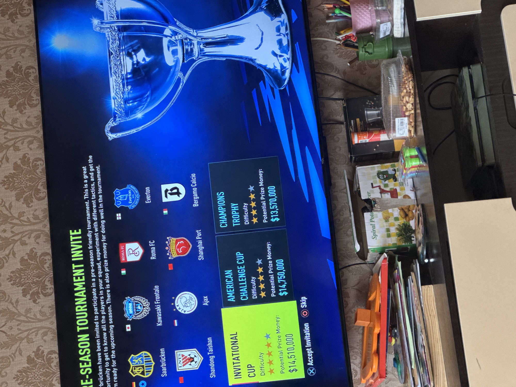 The image displays a television screen showing a video game interface, likely a football management simulation. The screen features a "PRE-SEASON TOURNAMENT INVITE" against a backdrop of a large, glowing trophy. The on-screen text explains that the tournament is an opportunity to experiment with tactics, get to know players, and earn prize money.
Three tournament options are available to the player:
1. **INVITATIONAL CUP**: Rated three stars difficulty, with a potential prize of $14,510,000.
2. **AMERICAN CHALLENGE CUP**: Rated five stars difficulty, with a potential prize of $14,750,000.
3. **CHAMPIONS TROPHY**: Rated two stars difficulty, with a potential prize of $13,570,000.
Logos and names of participating football clubs are visible, including Saarbrücken, Kawasaki Frontale, Shandong Taishan, Ajax, Roma FC, Shanghai Port, Everton, and Bergamo Calcio. The player can choose to "X Accept Invitation" or "O Skip".
Adjacent to the TCL television on the right, a dark brown shelf or table holds various household items. These include a clear plastic container of nuts or peanuts, colored pencils, a pink spool of yarn, a green military-style container, a box of "Spiral Potato" chips, a "BERG" branded item, a black and silver coffee grinder/mixer, and a dark green game console. Cables, books, and an orange toy are also scattered on the shelf. The background wall features a patterned, light brown wallpaper.