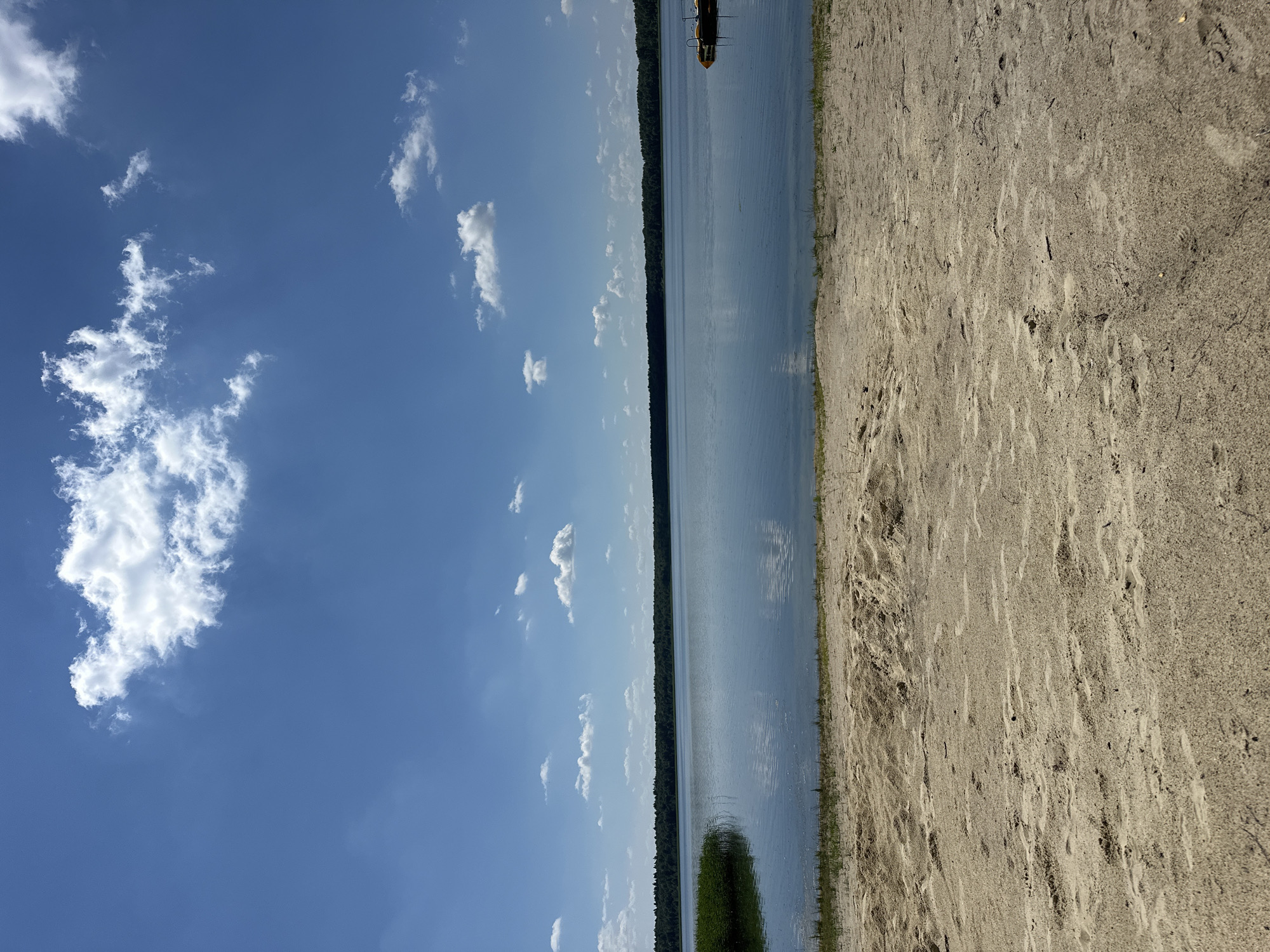 The image shows a sandy beach in Duvankul', Russia, with a calm lake in the background. The sky is blue with scattered white clouds. The lake is surrounded by a dense green forest. On the right side of the lake, a yellow floating platform is visible. The sand in the foreground is light brown and appears dry, with some small footprints and natural debris. The scene depicts a serene natural landscape on a clear day.