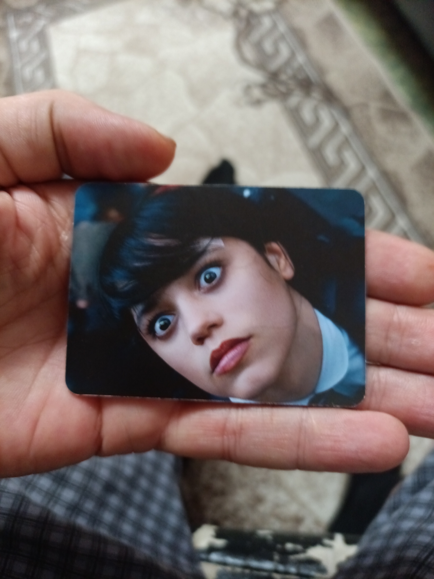 A person's hand holds a rectangular trading card featuring a close-up portrait of a young woman with wide, surprised eyes, dark hair, and red lipstick. The background of the card is dark and out of focus. The hand is resting on a patterned surface that appears to be a rug or carpet. The overall scene is an indoor setting. There is no indication of the city of Nyagan, Russia.