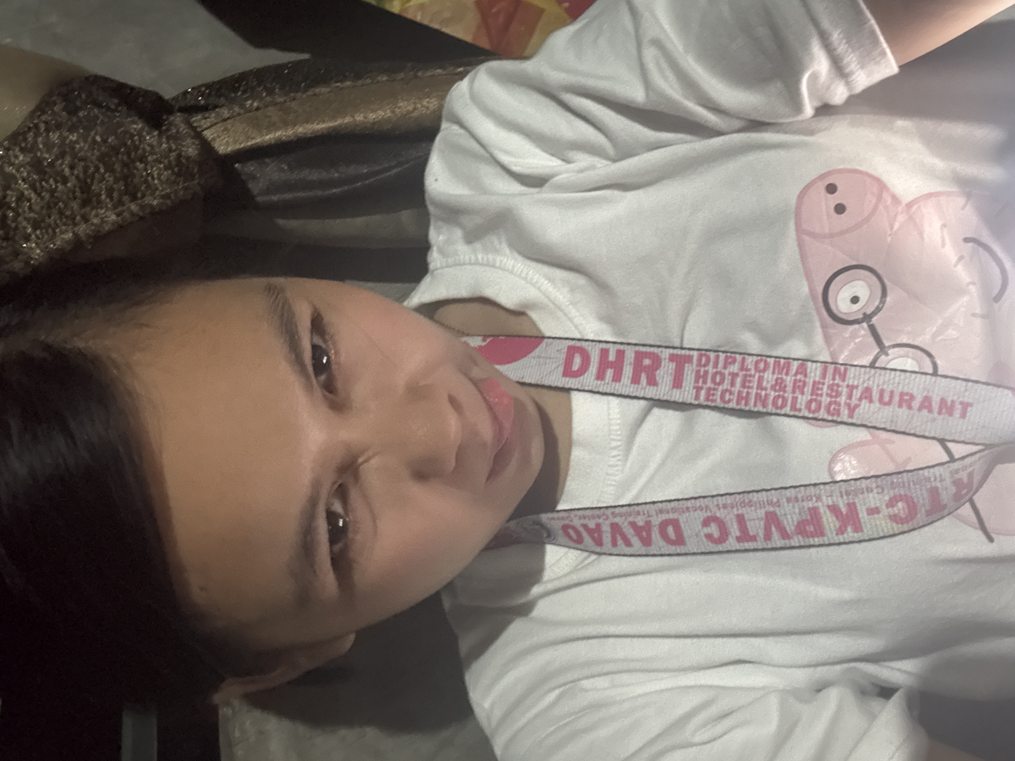 The image is a close-up, front-facing selfie of a young woman with dark hair, wearing a white t-shirt. She is making a playful, squished-face expression with her mouth pursed and her tongue slightly extended. A lanyard with text is visible around her neck, and it appears to say "DAVAO" on one side and "DHRT DIPLOMA IN HOTEL & RESTAURANT TECHNOLOGY" on the other, indicating the photo was likely taken in Davao City, Philippines, possibly at the Davao Region Technical Vocational College (DRTVC) or a related institution. A graphic of a cartoon pig character is printed on her t-shirt. The background is dimly lit and appears to be indoors, possibly a car or room, with some textured fabric visible behind her.