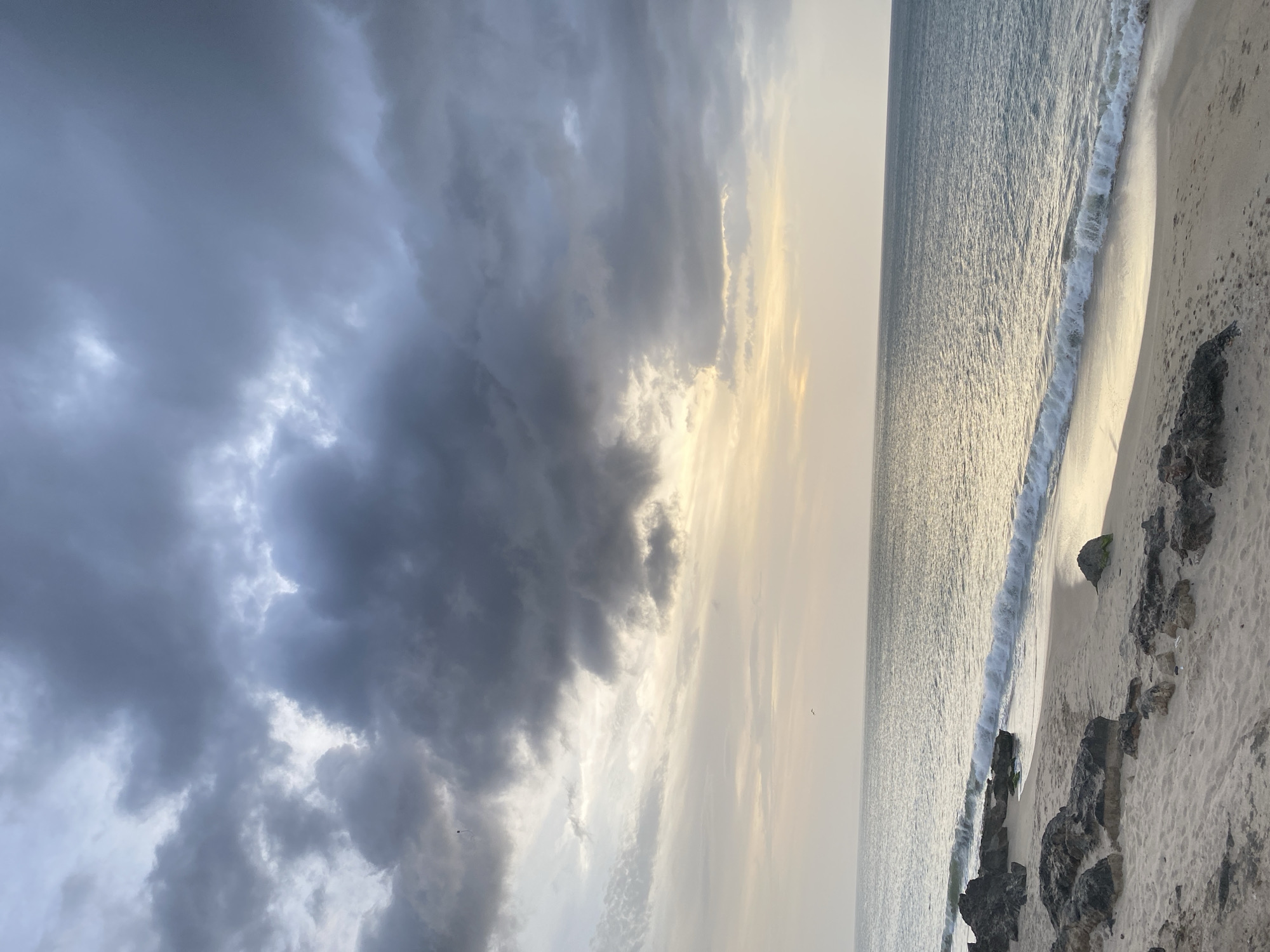 The image shows a seascape with a beach in Juan Griego, Venezuela. The left side of the image is dominated by dark, heavy clouds, while the right side shows the ocean meeting the shore. Gentle waves are visible at the shoreline. The beach is light-colored sand with dark rocks scattered along the edge where the water meets the sand. The overall lighting suggests either dawn or dusk, with the sky appearing overcast. There are no people visible in the image.
