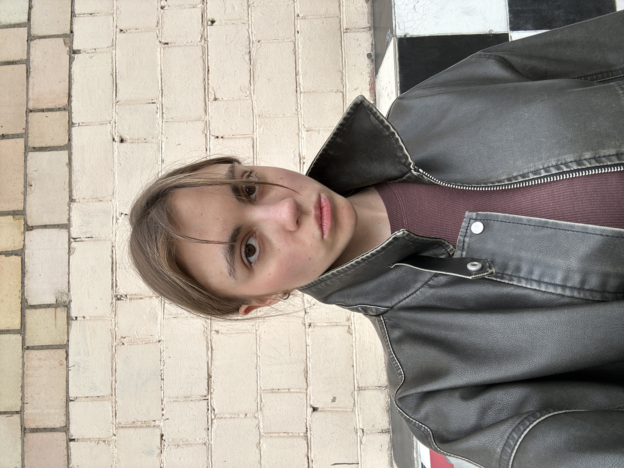A young woman with brown hair pulled back poses for a selfie in front of a brick wall. She is wearing a dark brown leather jacket over a burgundy long-sleeved shirt. The brick wall behind her is made of cream-colored bricks with visible mortar lines. To the lower right of the frame, a checkered pattern of black and white tiles is visible. The overall scene suggests an urban outdoor setting.