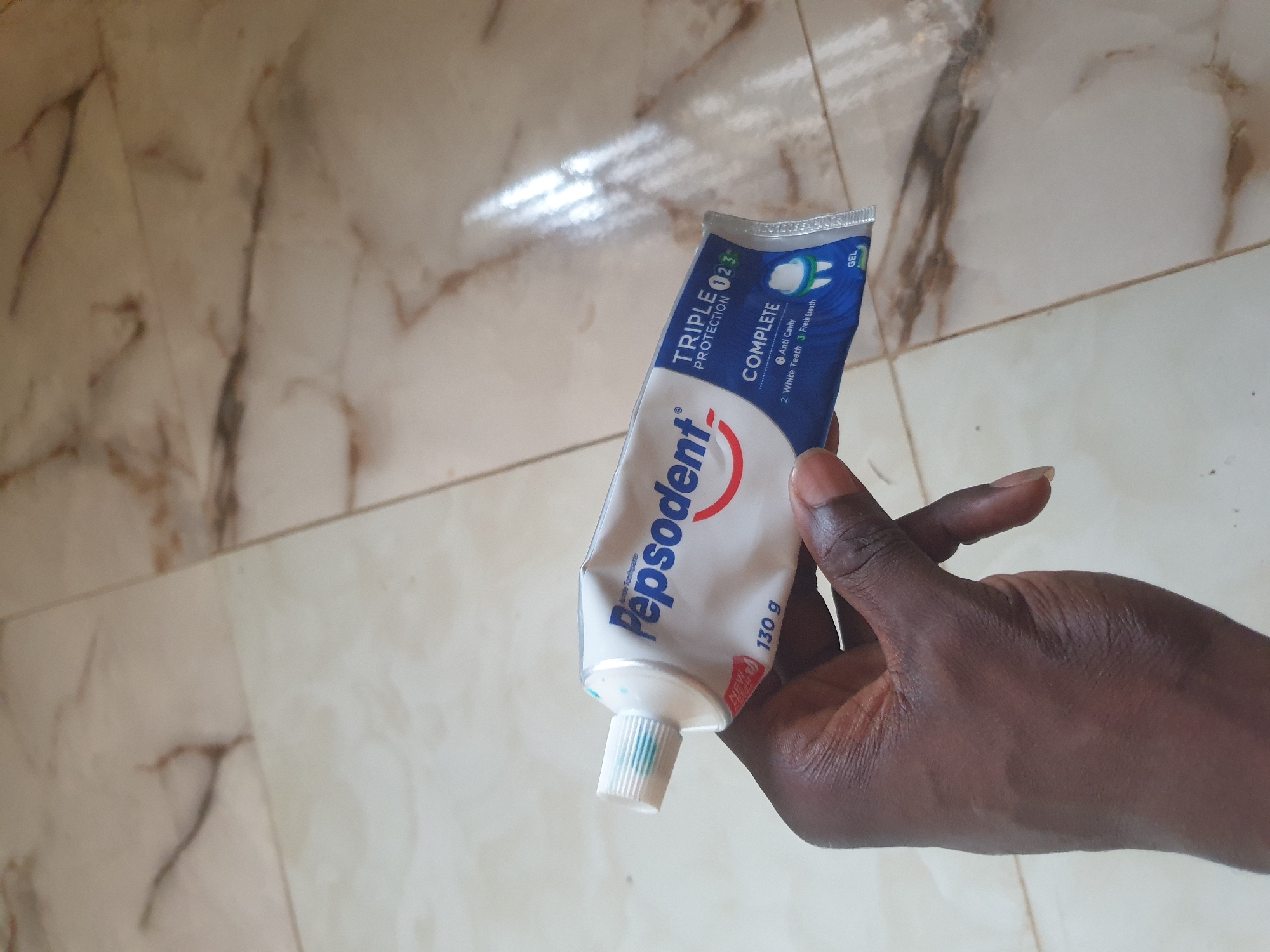 A person's hand holds a tube of Pepsodent Complete Triple Protection toothpaste. The toothpaste tube is white with blue and red lettering. The tube is partially squeezed, indicating recent use. The hand appears to be of African descent. The background consists of a tiled floor with a marble-like pattern in shades of beige and white, with brown veining. The lighting suggests an indoor environment, possibly a home or bathroom. The scene does not provide any specific indicators for the city of Anilelerin, Nigeria.