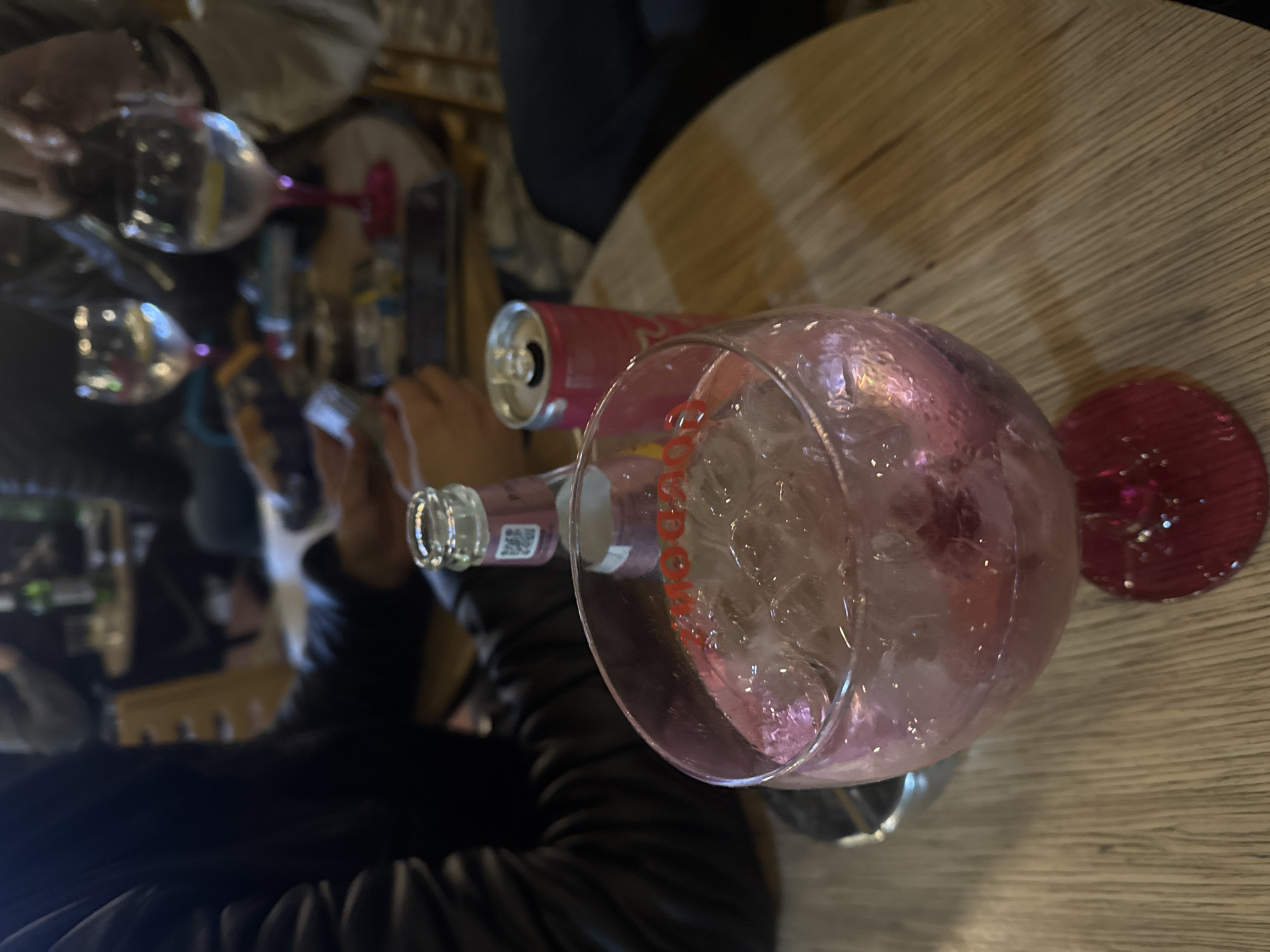 The image depicts a tabletop scene, likely in a restaurant or bar. A large wine glass filled with ice dominates the foreground, with a can of soda and a bottle of pink-colored liquid nearby. A person wearing a black jacket is visible in the bottom left corner. In the background, other people, glasses, and tableware are partially visible. The tabletop has a light wood grain pattern. It's impossible to determine the location as Plovdiv, Bulgaria, from the given image alone.