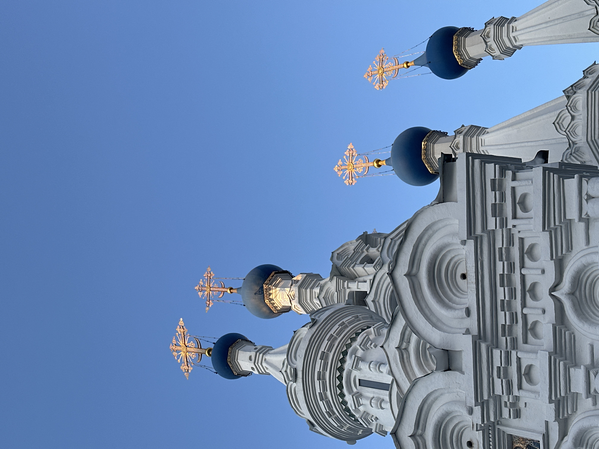 The image shows a low-angle view of the domes and architectural details of the Church of the Intercession at Fili in Moscow, Russia. Four blue onion domes are visible, each topped with a golden Orthodox cross. The church's white stone facade is intricately decorated with concentric circles, columns, and various geometric patterns. The sky is clear and blue. There are no people present in the image.