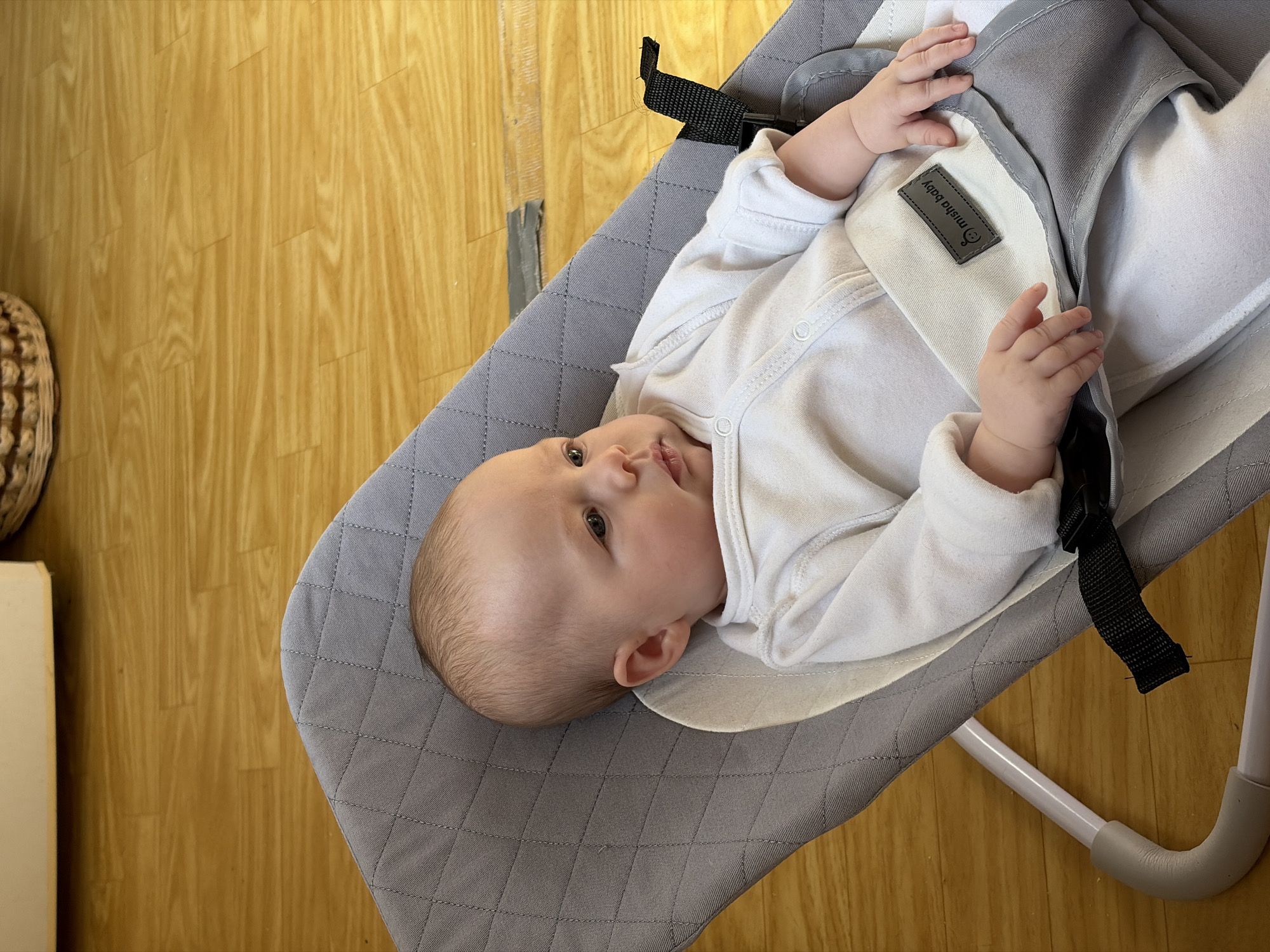 A baby lies in a gray baby bouncer. The baby is wearing a long-sleeved white onesie. The bouncer has black straps and a white fabric insert with a gray tag that says "Misha Baby." The bouncer has a light gray frame. The floor is light brown wood. A wicker basket and a tan cupboard are visible to the left. There is a piece of silver duct tape on the floor.
The location in the city of Chaikovsky, Russia is not possible to determine from the image.