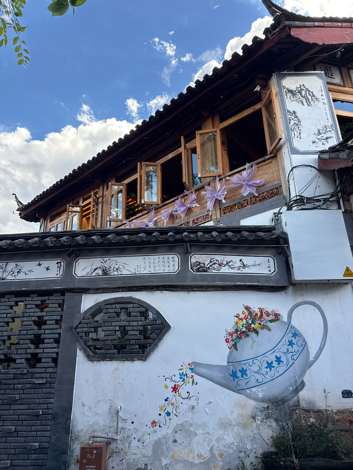 In Gucheng District, China, a traditional wooden building with a dark tiled roof is seen under a blue sky with white clouds. The building features multiple open windows, revealing interior lights. Decorative purple butterfly-like ornaments are attached to the building's facade. Below the wooden structure, a grey tiled wall displays traditional Chinese paintings of landscapes and birds. To the left, a section of a dark brick wall with a patterned opening is visible. The foreground is dominated by a white wall adorned with a mural of a watering can overflowing with colorful flowers. The watering can is depicted in grey and blue, with intricate floral patterns. Small, scattered flowers are painted as if spilling from the spout, creating a whimsical effect. The wall shows signs of wear and peeling paint, especially around the mural. Electrical conduits and a white electrical box are mounted on the wall to the right of the paintings. A small, rusty metal box is visible at the base of the wall, below the mural. The overall scene captures a blend of traditional architecture, artistic expression, and a hint of urban utility.