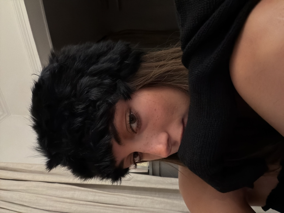 A woman with brown hair and freckles is wearing a large, fuzzy black hat. She's also wearing a black knitted garment. Her face is angled toward the camera, and she appears to be looking down. The background is out of focus, but beige curtains and the corner of a door frame are visible. The overall lighting is soft. The image is rotated.