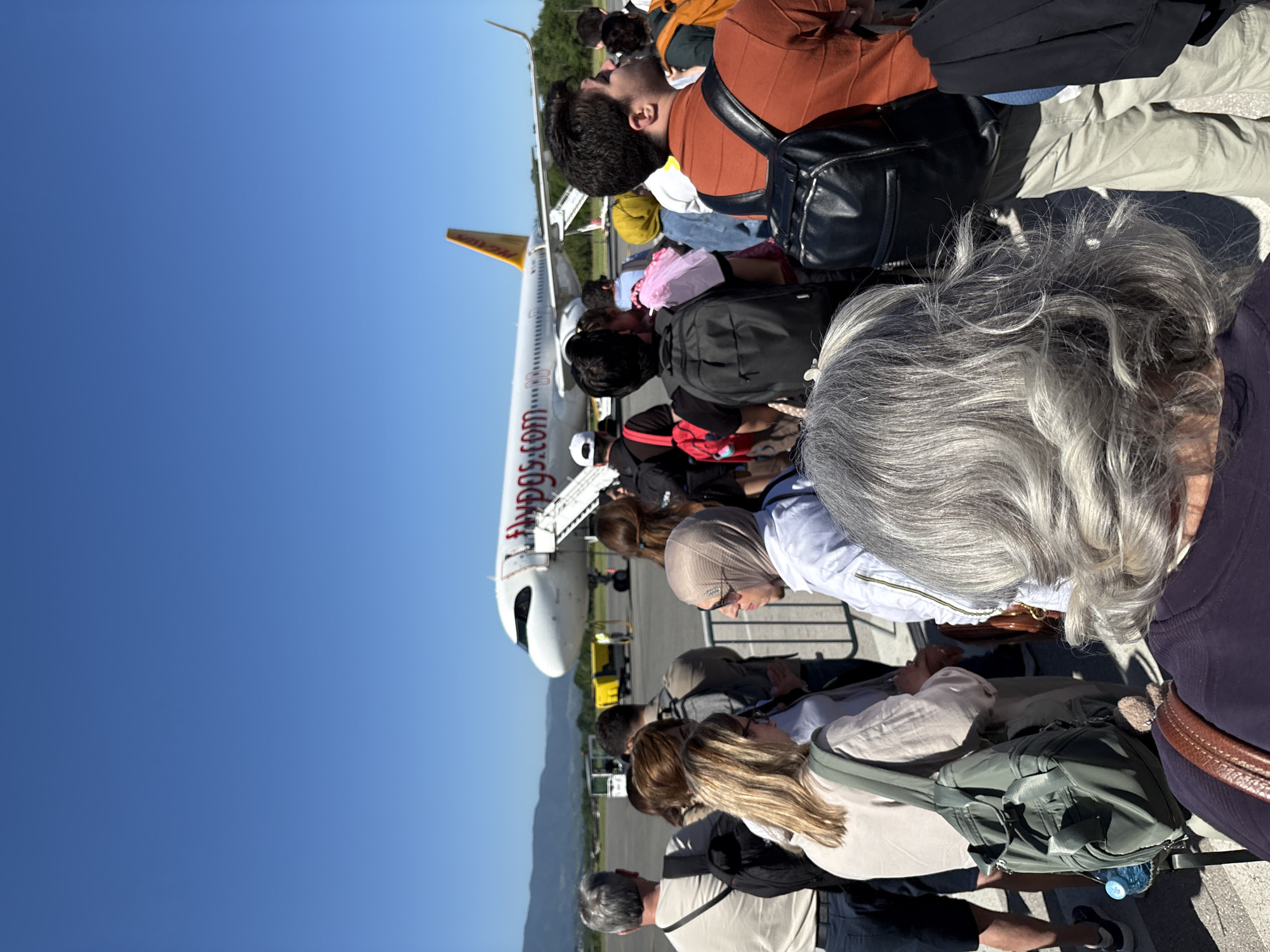 Passengers are queuing to board a Pegasus Airlines aircraft at an airport. The scene is outdoors on a sunny day with a clear blue sky. The location is likely the airport serving Gornje Dubrave, Bosnia and Herzegovina. People are seen with backpacks and other luggage. Some passengers are boarding the plane via stairs. The aircraft has "flypgs.com" printed on the fuselage.