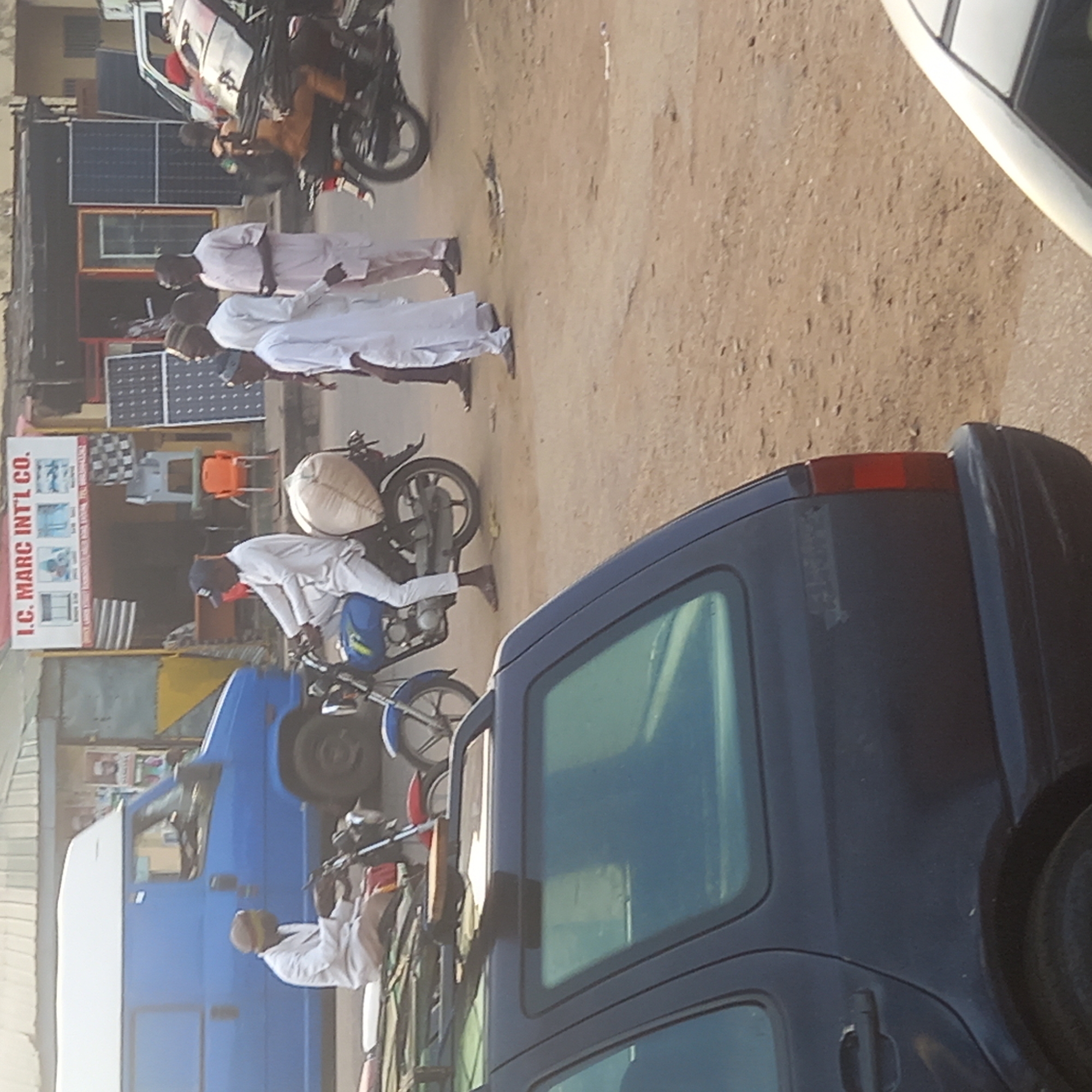 The image depicts a street scene in what appears to be Sardauna, Nigeria, based on visual cues. Several men dressed in traditional white attire are present. Some are walking along the side of the road, while others are riding motorcycles. One motorcycle has a large, white bag tied to the back. Several vehicles are visible, including a dark blue car, a light blue van, and other cars partially obscured by the foreground objects. A building on the left side of the frame features a sign that reads "I.C. MARC INTL CO.". Solar panels are displayed in front of the building. The ground appears to be a dirt road or unpaved surface.