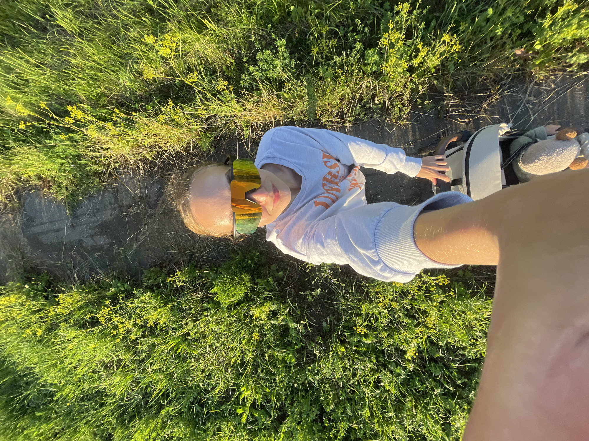 A woman is taking a selfie in a grassy area with a dirt path. She is wearing a white sweatshirt and reflective sunglasses. She is holding a bag with a stuffed animal attached to it. The sun is shining, casting long shadows. The background is filled with green grass and small yellow flowers.
