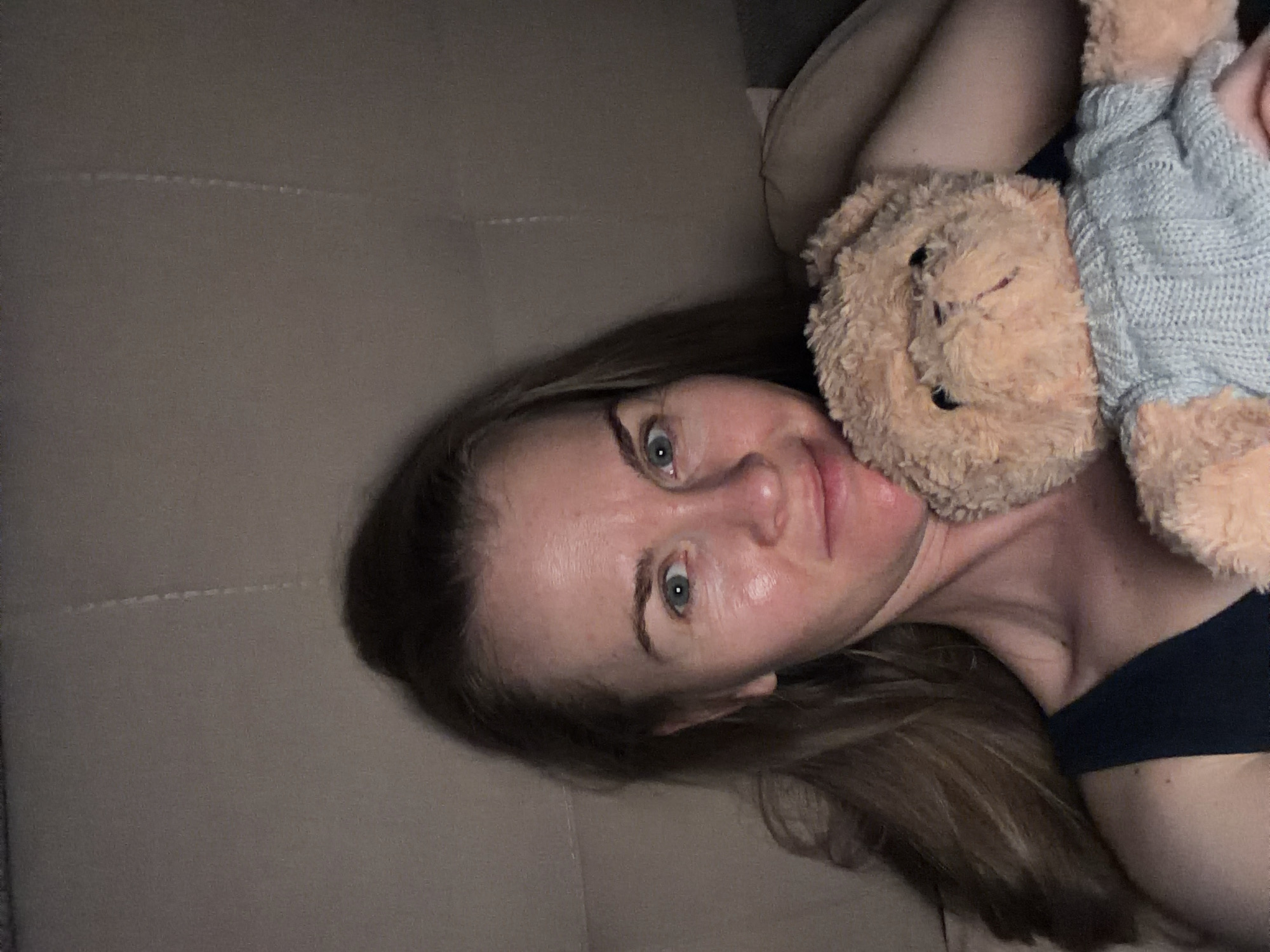 A woman, with long brown hair and blue eyes, is holding a teddy bear. She is wearing a dark-colored tank top. The teddy bear is light brown and wearing a light blue knitted sweater. The background is a textured surface, possibly a couch or wall. The lighting is dim and appears to be artificial. There is no indication of location in the city of Khabarovsk, Russia.