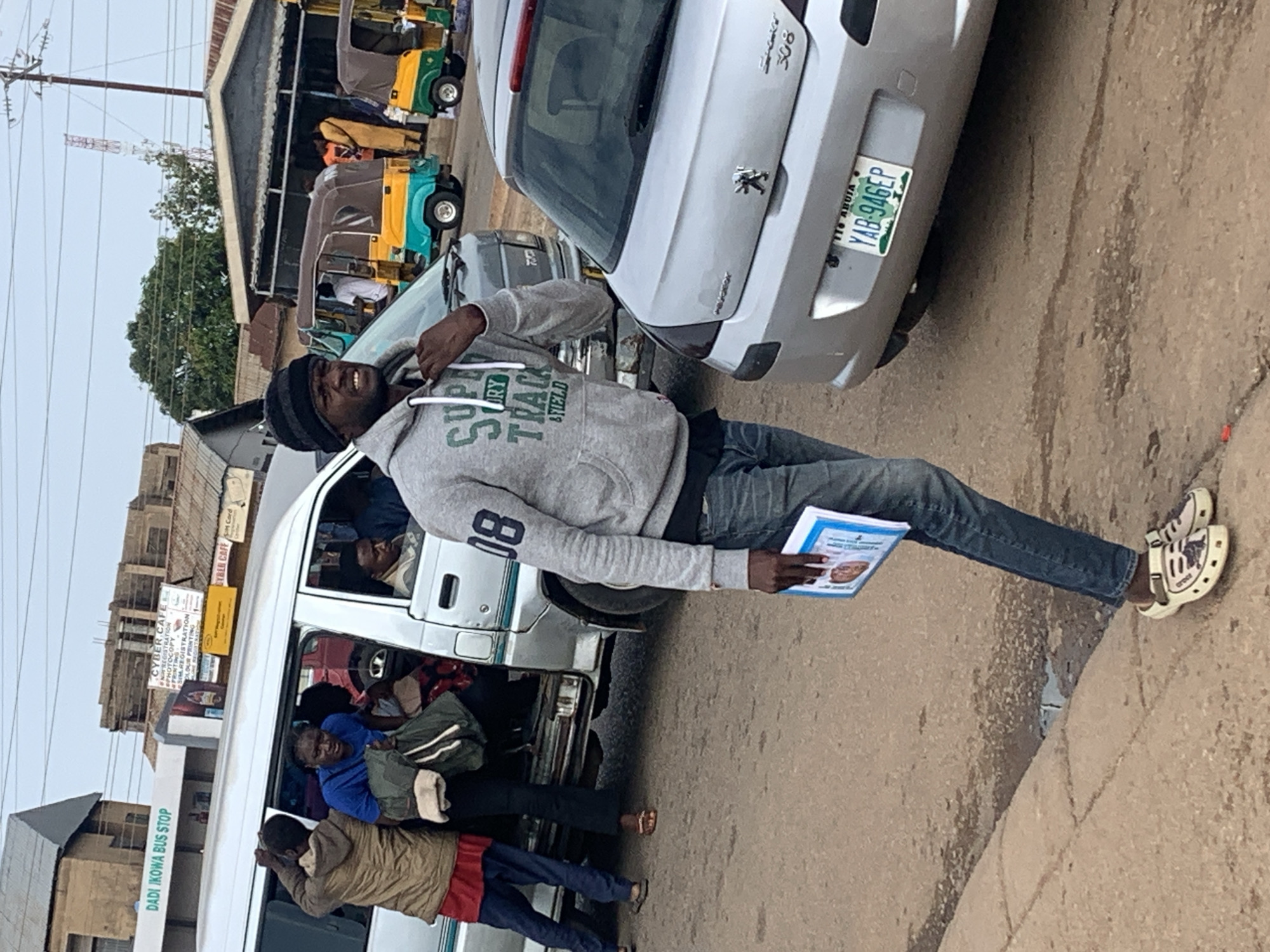 The image depicts a street scene in Gura Top, Nigeria. A man wearing a grey hoodie, blue jeans, and white crocs is the central figure. He is holding a booklet with a picture of a person on it. To his left, a silver Peugeot 308 car is parked. Behind the car, a silver minivan with passengers inside is stopped, and several people are interacting around its open doors. Further in the background, three colorful auto-rickshaws are visible, and beyond them, buildings and a utility pole with antennas can be seen. The ground is paved with asphalt and shows some signs of wear and moisture. The overall lighting suggests it is daytime.