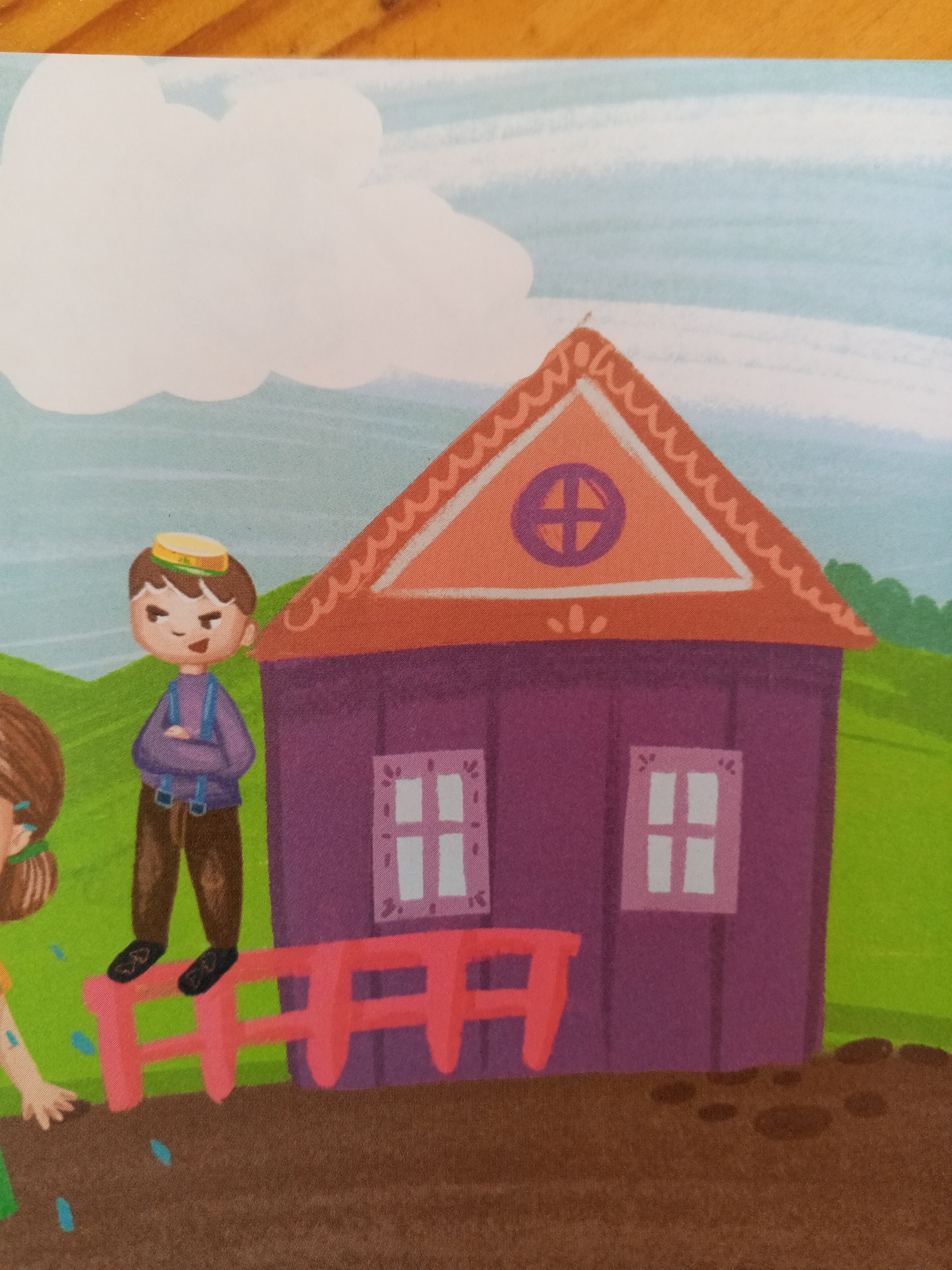 The image depicts a cartoon illustration of a rural scene. In the foreground, a young boy with brown hair, wearing a yellow hat, purple shirt, brown pants, and black shoes, stands with his arms crossed on a red fence. He has a stern expression. To his left, a partial view of a young girl with brown hair and a green ribbon is visible, reaching out her hand towards the bottom of the frame. A purple house with an orange roof and white-paned windows stands in the background, flanked by green hills and a pale blue sky with a white cloud. The ground is depicted as brown soil with some small dark spots. There is no indication of the city of Chistopol, Russia, in the image.