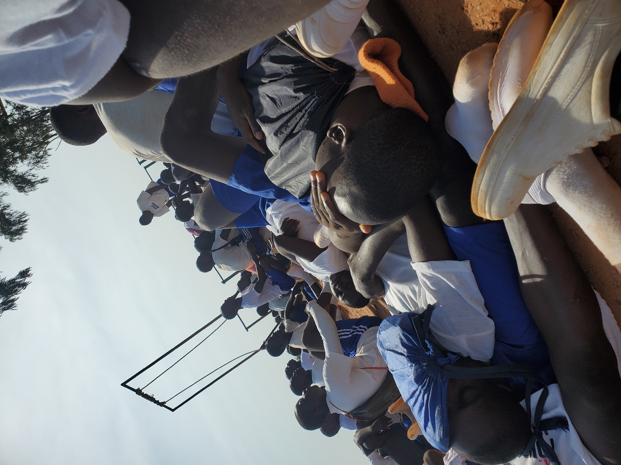 In Barakin Rafin Gora, Nigeria, a group of young males, likely students, are seen lying down on an uneven, sandy surface under a bright, overcast sky. They appear to be resting or waiting, with some dressed in white shirts and blue shorts, while others are in similar attire. A black drawstring bag is placed under one individual's head. In the background, a metal structure, possibly a swing set or part of a sports facility, is visible against the sky, with trees at the top of the frame. The overall scene suggests a communal gathering in an outdoor, possibly school or recreational, setting.