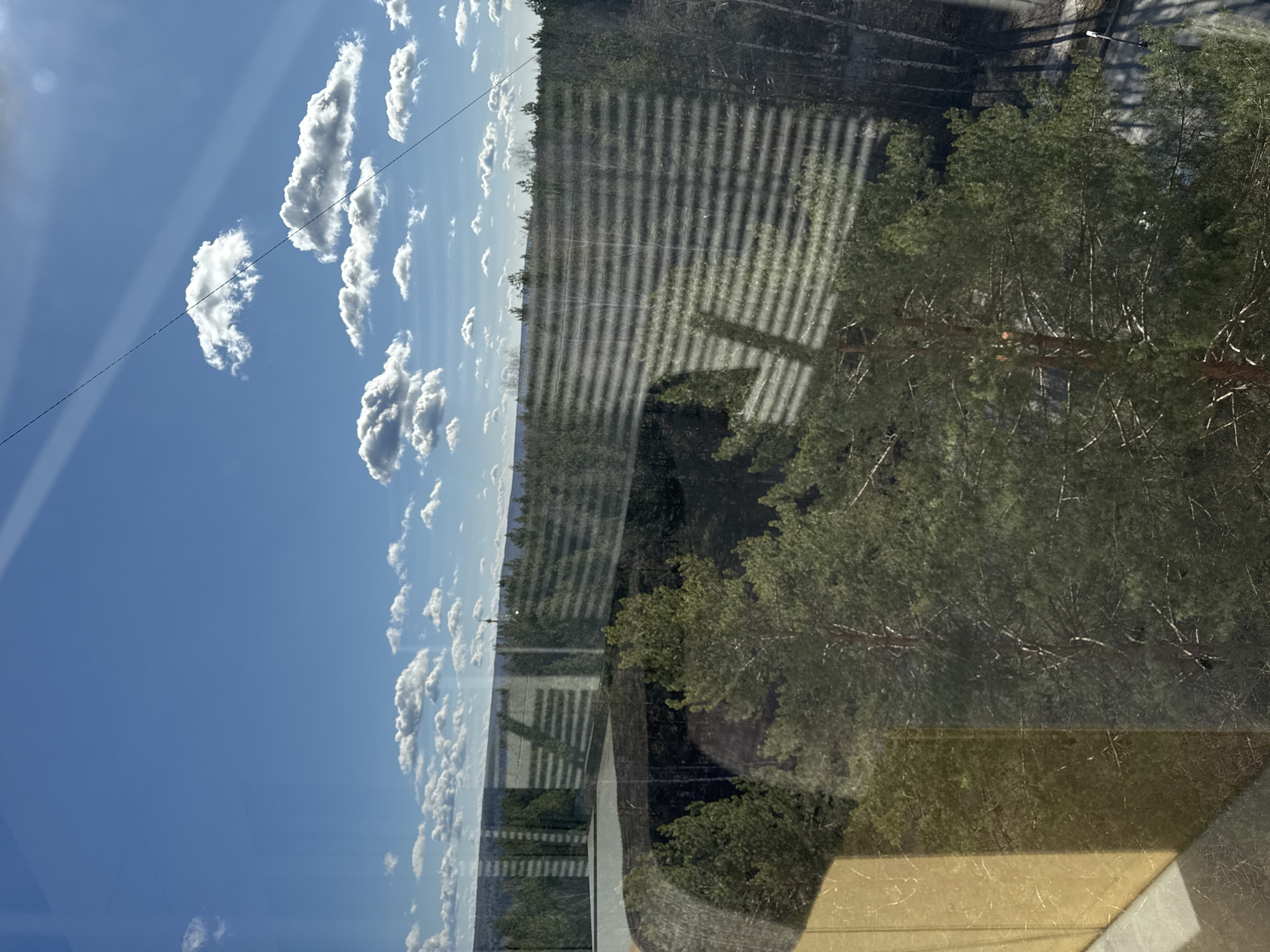 The image shows a view from a window, likely in a building located in the city of Chaikovsky, Russia. The reflection in the glass partially obscures the outside scene.
The upper left of the image reveals a clear blue sky with scattered white cumulus clouds. A power line is visible across the top left portion.
Beyond the reflection, the scene depicts a forested area with trees. A section of what appears to be a road or pathway runs through the trees. Overlaid on the scene are shadows of what appear to be window blinds. The window is near a building, where we see a section of siding.