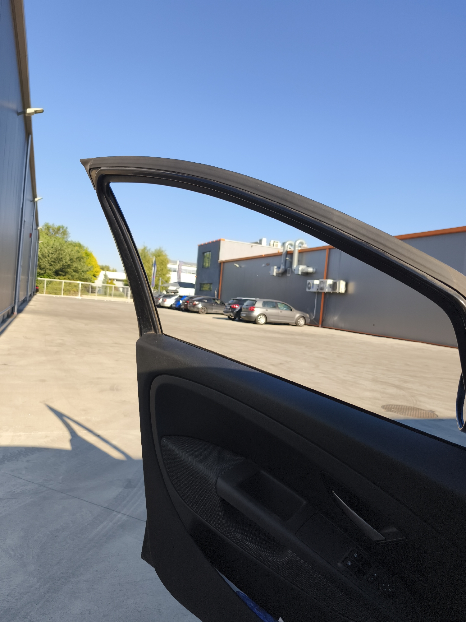 The image, taken from inside a car with the driver's door open, looks out onto a paved lot in front of a modern building. The sky is a clear, bright blue. To the left, the side of the building is visible, with lights mounted on its exterior. In the distance, several cars are parked. The building in the background is a two-story structure with a gray facade and orange trim around the windows. External pipes and air conditioning units are visible on the building's exterior. The car door interior shows a black armrest with controls for the windows and a door handle. The shadow of the open car door and a portion of the car's interior are cast on the concrete ground.