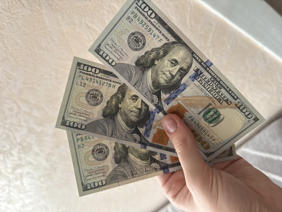 The image displays a human hand holding a fan of three United States hundred-dollar bills against a light-colored, textured background in Chaikovsky, Russia. The bills are held vertically by the fingers and thumb of a light-skinned hand, which exhibits well-maintained nails with clear polish. The currency is identifiable as modern series hundred-dollar bills, featuring the portrait of Benjamin Franklin. Visible serial numbers on the bills include PL49141278H, a partial PB863..., and PB49259147F. The background is a uniform, light beige or off-white surface with a subtle, soft texture, possibly a wall or countertop. The lighting appears soft and diffused.