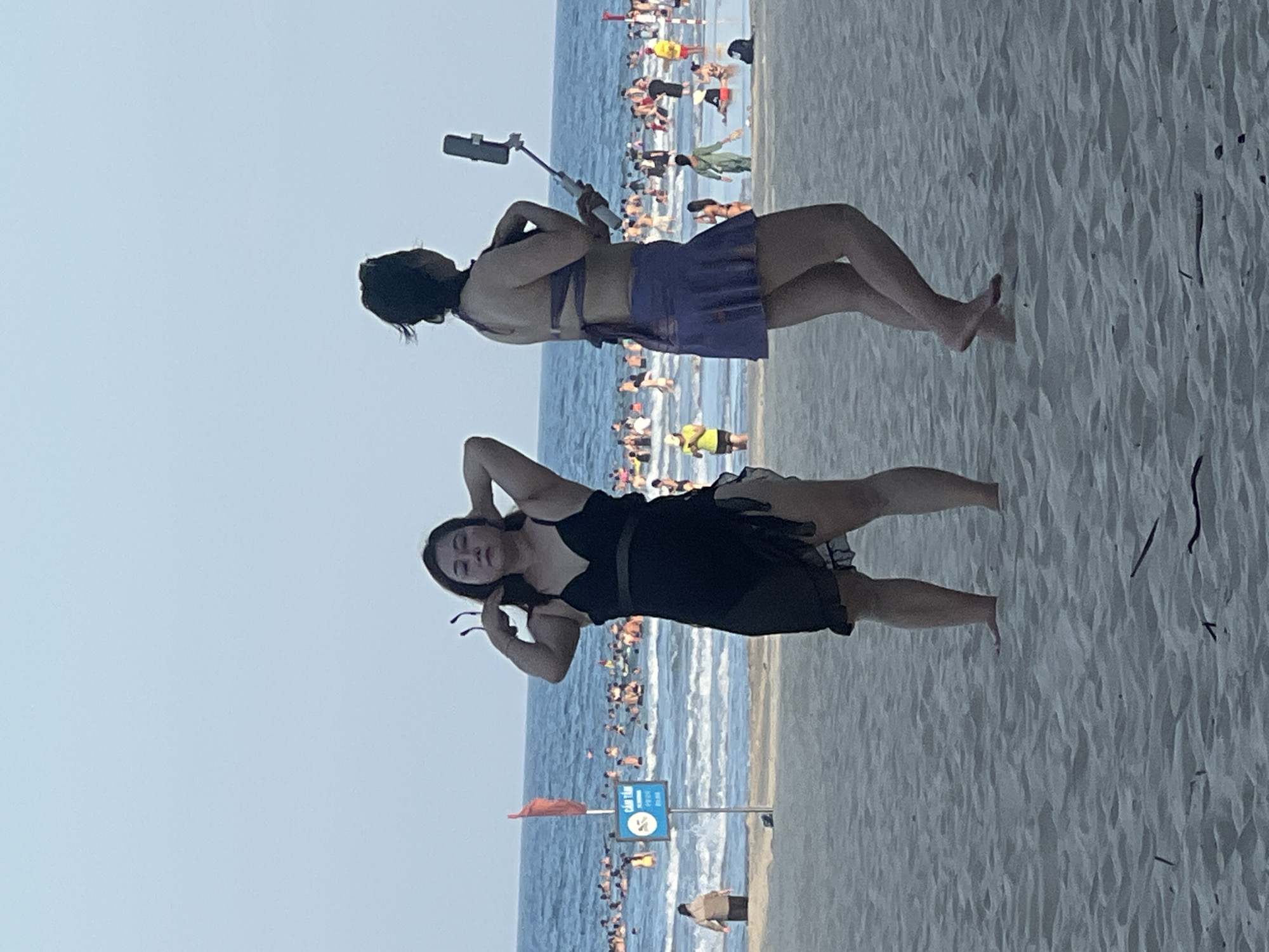 The image shows a beach scene at My Khe Beach in Da Nang, Vietnam. In the foreground, two women are standing on the sand. The woman on the left is wearing a black swimsuit and has her hands on her head. The woman on the right is wearing a purple bikini with a skirt and is holding a selfie stick with a phone, taking a picture of herself. In the background, the ocean is crowded with people swimming and enjoying the water. A red flag and a "Cam Tan" (No Swimming) sign are visible on a pole in the water. The sky is clear and the weather appears to be warm.