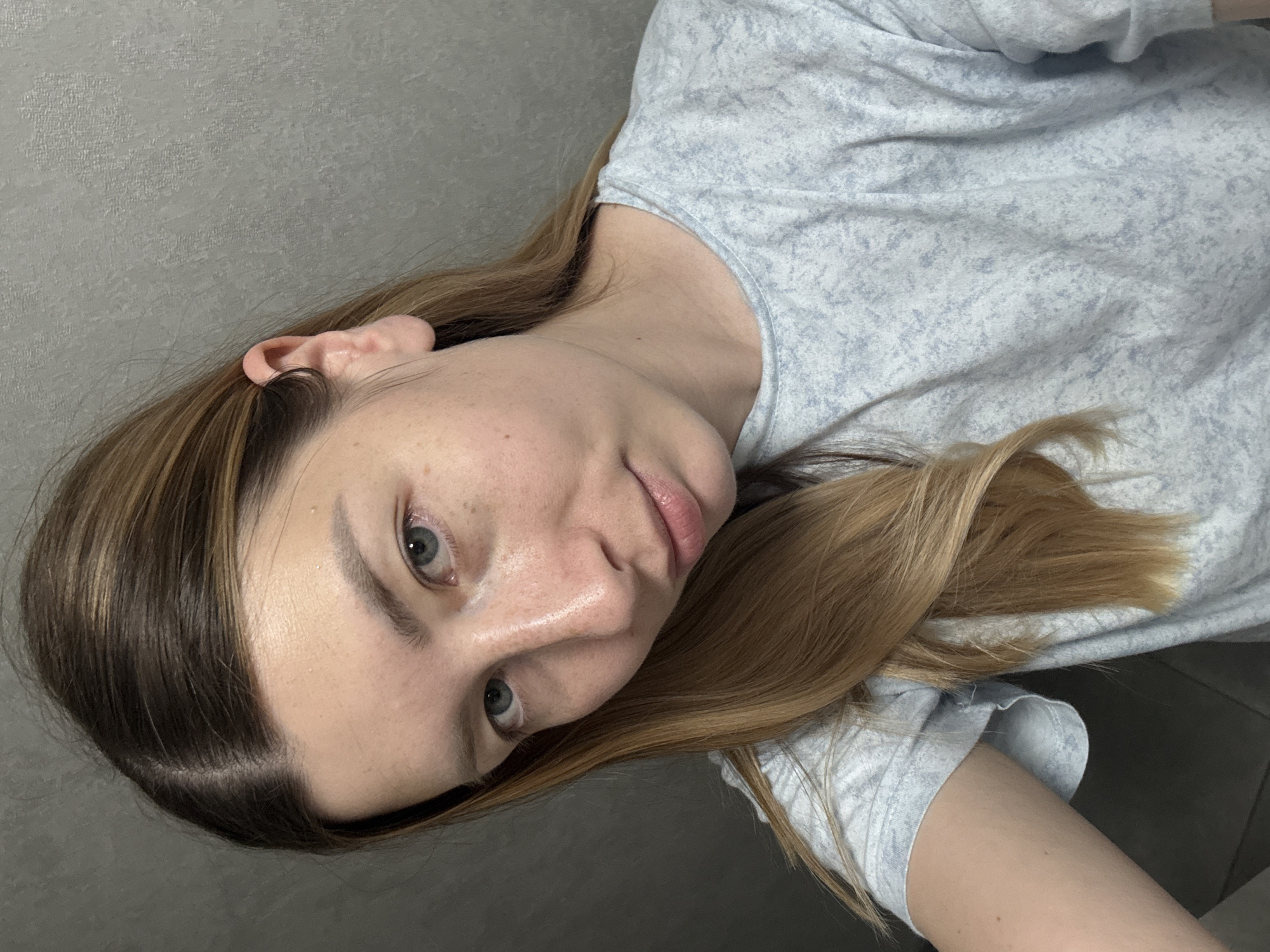 A woman is shown in a close-up portrait, appearing to be indoors. The background is a light grey wall. The woman has fair skin, freckles, and light eyes. Her hair is long, light brown with blonde highlights. She is wearing a light-colored t-shirt. No information is available to associate the image with Moscow, Russia.
