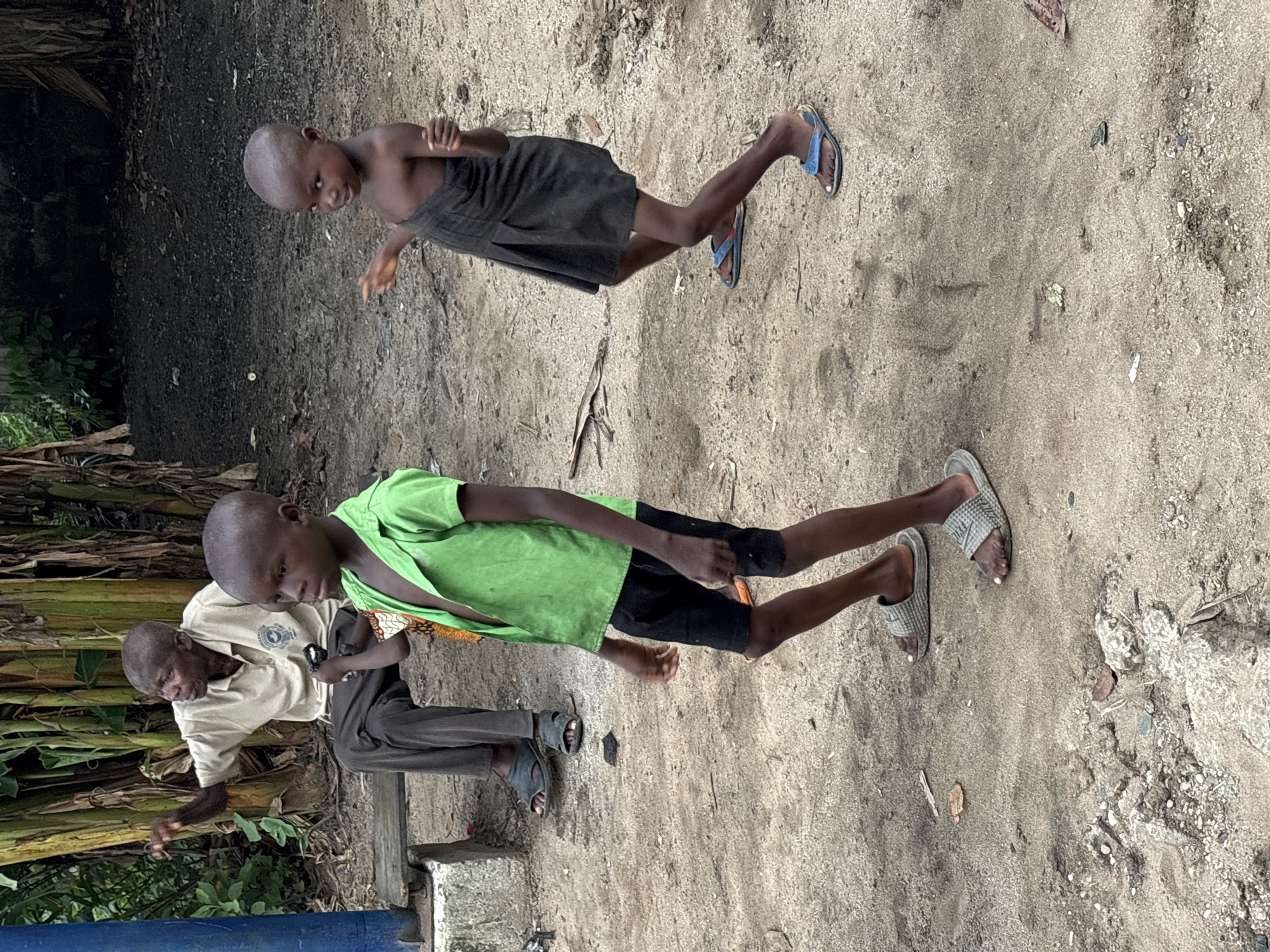 The image shows three individuals in an outdoor setting, likely in Ikot Ekpene, Nigeria, given the prompt. Two children are visible: one near the top of the frame wearing a dark skirt and blue flip-flops, and another in a green shirt and black shorts, wearing grey sandals. An adult male, wearing a light-colored shirt and dark pants, is also present. The ground appears to be sandy or dirt. A cluster of banana trees can be seen along the left side of the frame. The children appear to be in motion.