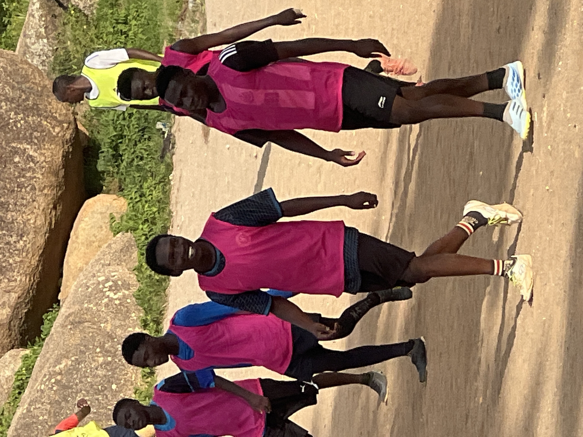 This image captures a group of young men, appearing to be in their late teens or early twenties, participating in an outdoor activity, possibly a sports training session, on a dusty, sun-drenched ground. They are all wearing athletic attire, with most of them sporting bright pink training vests over dark t-shirts and black shorts. Several individuals have on socks and athletic shoes, in various colors and designs. The background features large, weathered rocks and dense green foliage, indicative of a natural or semi-developed outdoor area. In the distance, two individuals are visible, one wearing a yellow training vest over a white t-shirt, and another in a yellow shirt. The lighting suggests it is daytime, with the sun casting shadows on the ground. The setting, with its distinctive rock formations and vegetation, is consistent with locations found in and around the city of Jos, Nigeria, known for its rocky terrain and highland climate.