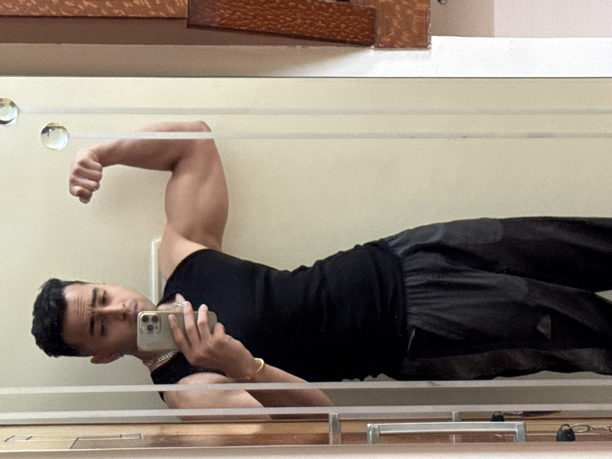 A person is posing in front of a mirror, taking a selfie, and flexing their bicep. The person is wearing a black tank top and black pants. The background appears to be a white or light-colored wall. There are decorative features above and below the mirror. It is not possible to determine if the image was taken in the city of Kolkata, India.