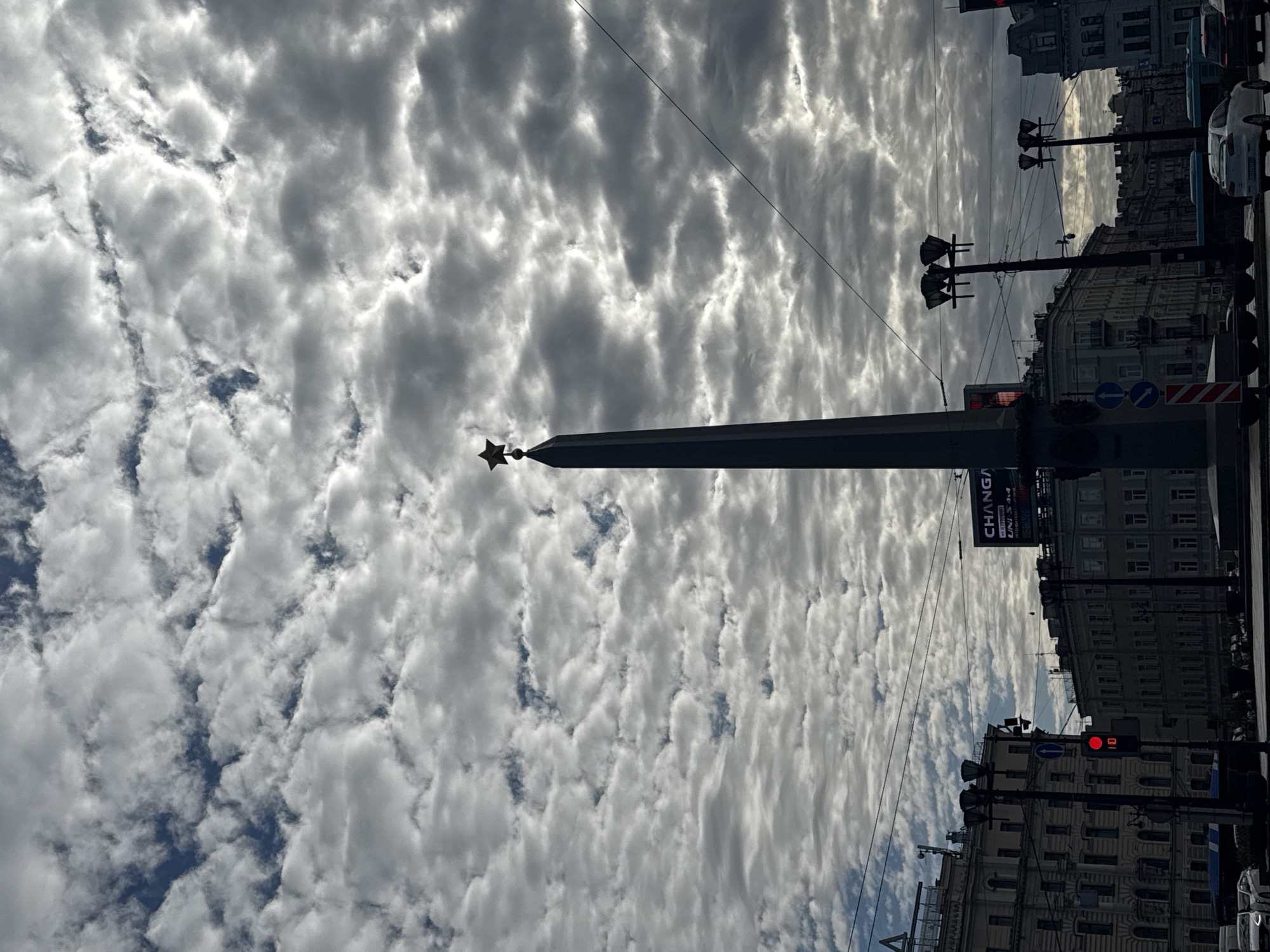 A street-level upward view captures an urban scene in Saint Petersburg, Russia, dominated by a tall monument against a dramatic sky. In the center, a dark, slender obelisk-like column rises, topped with a radiant, golden five-pointed star.
The sky above is filled with a dynamic display of textured, white and grey altocumulus or stratocumulus clouds, creating a rippled pattern with patches of brighter sky breaking through. The lighting suggests either a bright, cloudy day or the sun is subtly obscured by the cloud cover, casting an even, diffused light.
To the right of the obelisk, several multi-story buildings line what appears to be a street, their facades shadowed but showing architectural details. Numerous utility poles and streetlights extend upwards, crisscrossed by overhead wires. A traffic light prominently displays a red signal. Street signs are visible, including blue circular signs with white arrows indicating traffic flow, and a red-and-white striped sign.
A large sign or billboard is partially visible on one of the buildings, displaying the text "CHANGA", followed by smaller text "UNIS 3x4" and "KITCHEN". Several cars are parked or moving along the street next to the buildings.
The image captures a typical urban environment under a visually striking sky, with the monument serving as a focal point, characteristic of Saint Petersburg's historical and monumental landscape. The scene conveys a sense of everyday city life enhanced by the grandeur of the natural elements above.