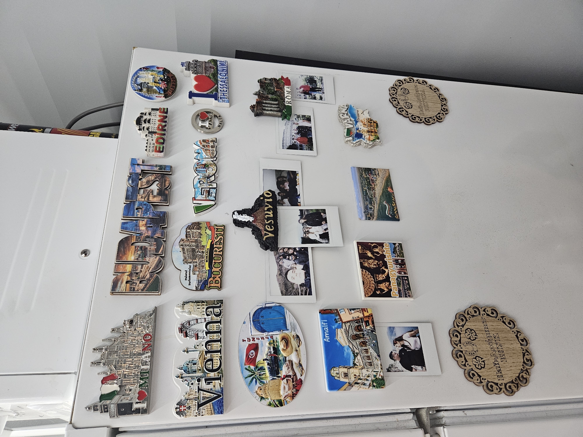 The image displays the front of a white refrigerator covered with a diverse collection of travel magnets and several instant photographs. The arrangement suggests a personal display of memories and souvenirs, likely in a home setting.
The majority of the items are refrigerator magnets, many of which are shaped and inscribed with names of cities and countries. Visible place names include "THESSALONIKE," "I ❤️ MALTA," "EDIRNE," "ROMA," "VESUVIO," "BUCURESTI" (Romania, with "Palatul Parlamentului"), "MILANO," "VIENNA," "TUNIS," "Amalfi," "Panagyurishte," and "SUNNY BEACH." One magnet, implied by the location context, also says "ELIN PELIN." These magnets showcase a range of architectural styles, flags, and natural landmarks.
Interspersed among the magnets are several small, square instant photographs featuring various individuals and groups, appearing to be friends or family, smiling and posing in different settings.
Two ornate, oval-shaped wooden decorative magnets are also present. One features a pattern with shamrocks and a horseshoe, while the other, in Bulgarian, reads "Най-добрият ВНУК" (Naĭ-dobriyat VNUK), meaning "The Best Grandson."
The overall scene is a vibrant and personal testament to travels and cherished moments, with the collection serving as a visual diary of experiences.