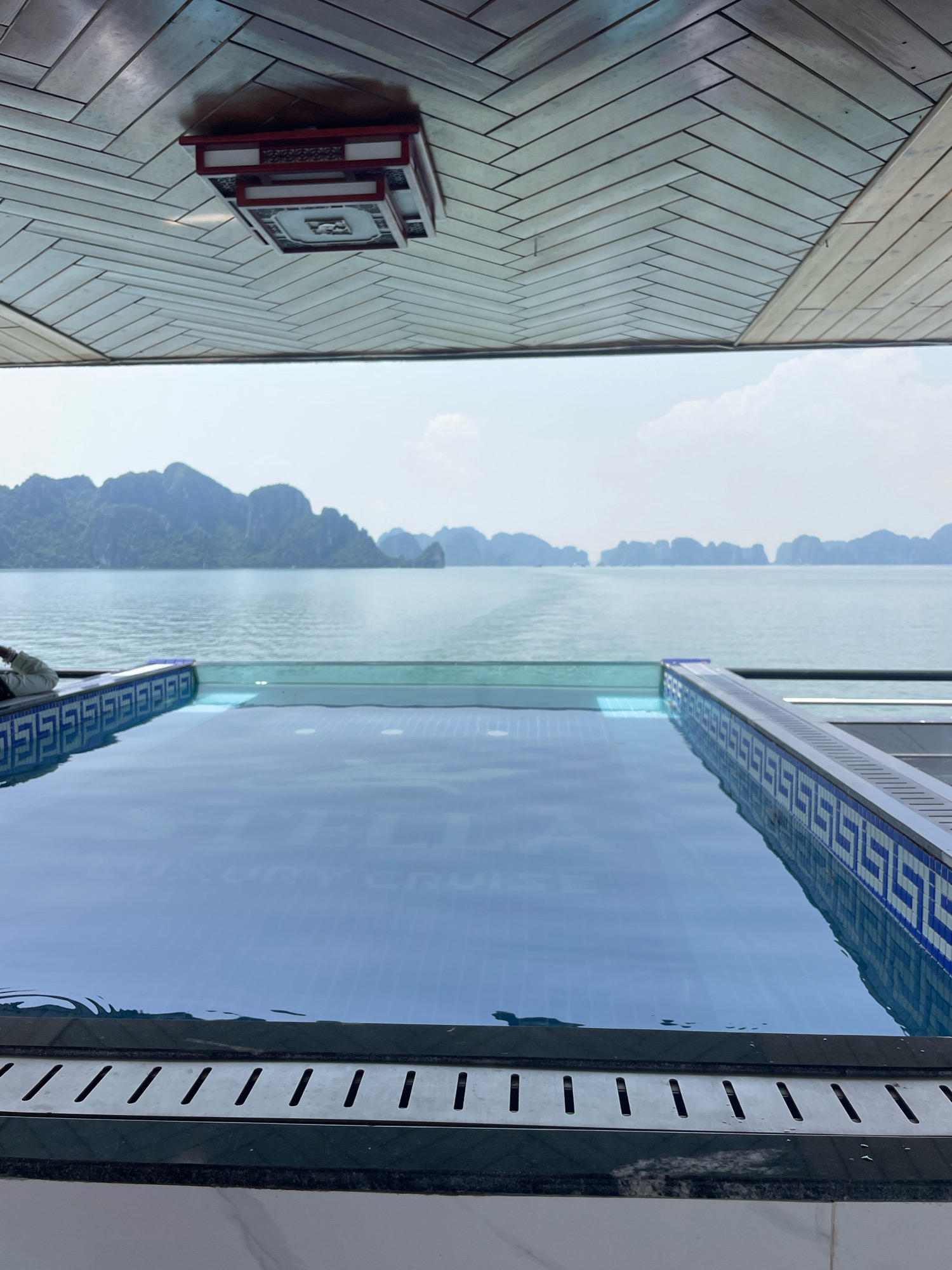 An infinity pool, filled with calm, reflective blue water, sits on the deck of a vessel. The pool is bordered by a decorative blue and white tiled edge on the left and right, with a sleek grey metal edge along the front. Faint text, possibly indicating "LUXURY CRUISE," is visible on the bottom of the pool. Beyond the pool, a vast expanse of calm, grey-blue water stretches to the horizon, where numerous lush, green islands rise dramatically from the sea. The sky above is a hazy white with sparse clouds. The interior of the vessel's deck is visible in the upper portion of the image, featuring a wooden ceiling with a herringbone pattern and an ornate, square, red and white light fixture. The scene is likely located in Ha Long Bay, Vietnam, known for its karst island formations and cruises.