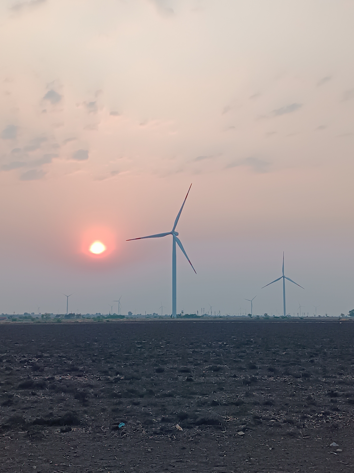 The scene depicts an expansive outdoor environment in Kottalapalli, India, observed during what appears to be either dawn or dusk. The foreground consists of a dark, uneven field of barren or tilled earth, presenting irregular textures and containing sparse debris, including a light blue plastic item visible towards the lower-center. A prominent, bright orange-red sun disc is situated in the left-center of the sky. Numerous three-bladed wind turbines are scattered across the distant horizon, forming a wind farm. One large white wind turbine with red-tipped blades stands notably in the mid-ground, positioned to the right of the sun. The sky above exhibits a color gradient, transitioning from vibrant orange and pink hues near the sun to lighter grays and purples towards the upper and right portions, with several scattered, indistinct clouds. No individuals, animals, or specific actions are discernible within the frame.