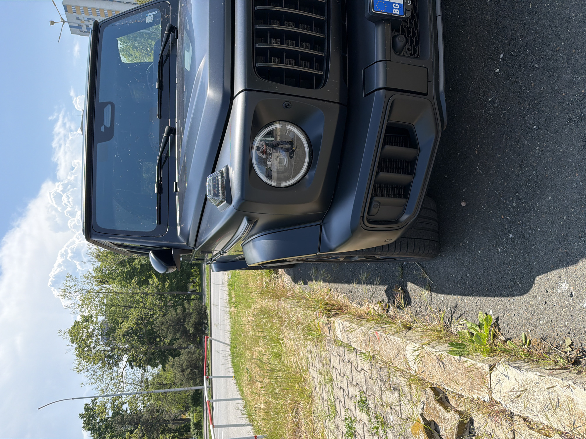 The image shows the front of a black SUV, specifically a Mercedes-Benz G-Class (G-Wagon), parked on the side of a paved road. The car is positioned with its front end facing the viewer and slightly to the right. The license plate is partially visible, displaying "BG," the country code for Bulgaria. The car appears to be in relatively good condition.
The surrounding environment consists of vegetation (grass and trees) along the roadside. A low retaining wall constructed of interlocking pavers separates the road from the vegetation. A light pole is also visible in the background. The sky is blue with some clouds. Based on the license plate code and general appearance, the location is likely in an urban area in Burgas, Bulgaria, but without specific landmarks, a precise location is difficult to determine.